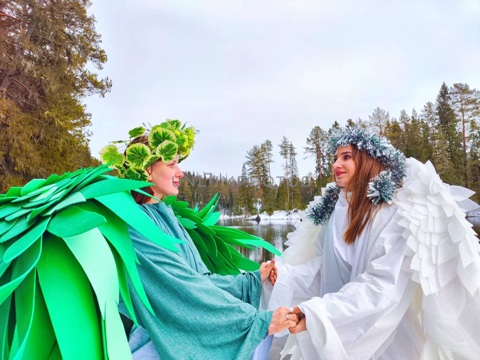 Two girls in vibrant green and white dresses embrace nature while role-playing the enchanting meeting of winter and spring by a serene lake.