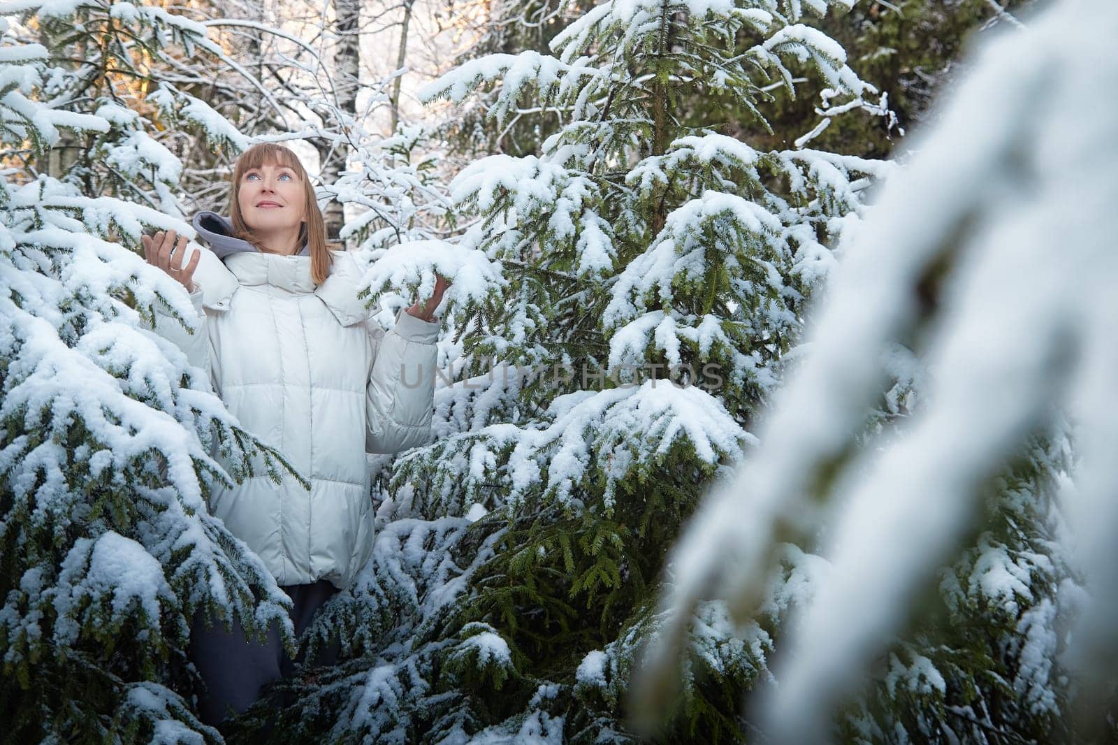A middle-aged woman walks through a snowy forest, surrounded by snow-laden trees, embracing the beauty of winter during the festive season.