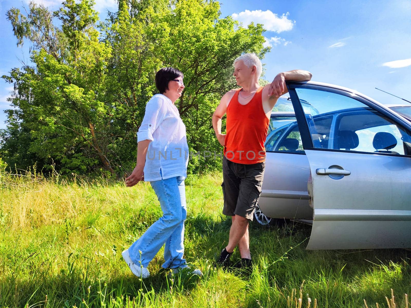 A middle-aged couple embraces the warmth of a sunny day while standing beside their parked car. They are laughing and sharing joyful moments in a lush green area, radiating happiness and connection.