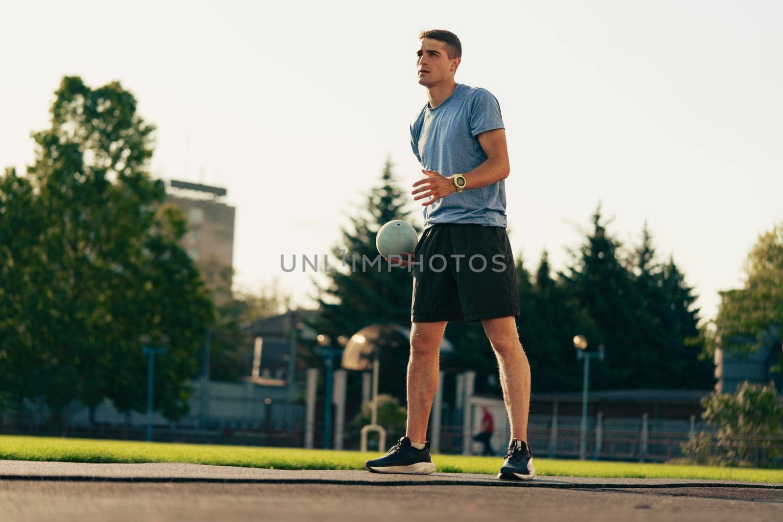 A young man stands on a grassy field, holding a soccer ball, wearing athletic clothing. It is a bright day, with trees and buildings in the background.