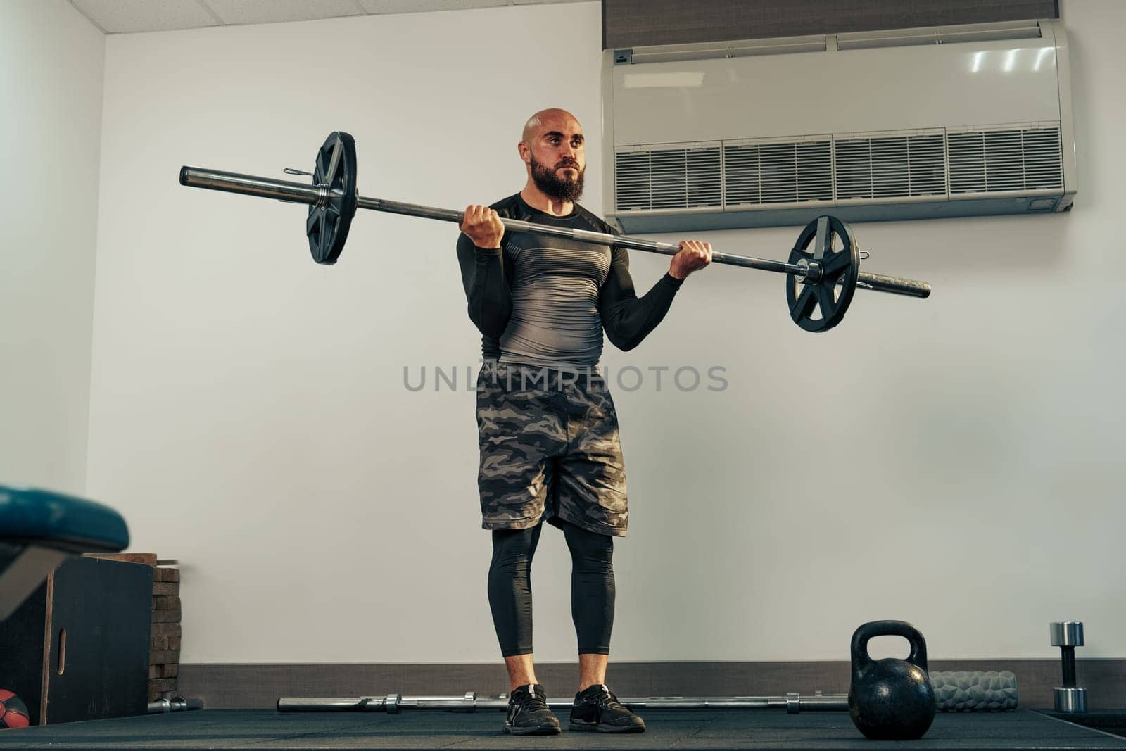 A man engaged in strength training holds a barbell at shoulder height in a gym. He focuses on his form while surrounded by gym equipment and weights.