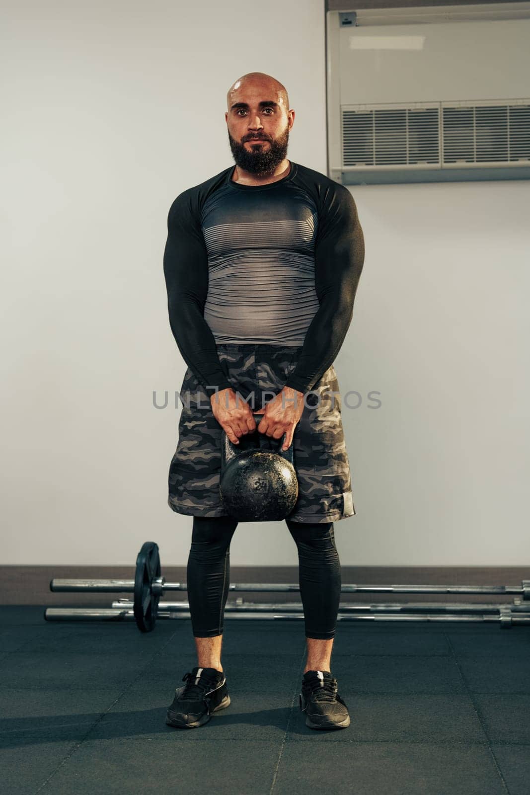 A fit man stands in a gym holding a kettlebell, wearing a fitted long-sleeve shirt and shorts. The gym features various exercise equipment and weights, indicating an active workout environment.