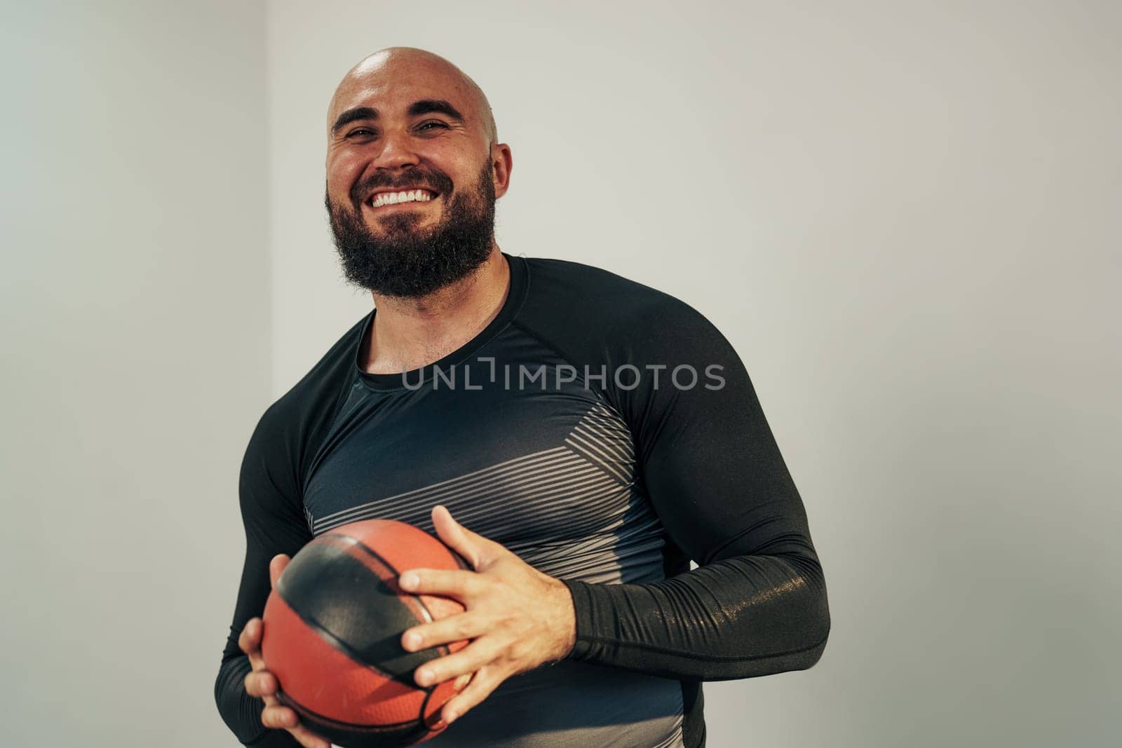 A man with a beard smiles while holding a basketball indoors. He wears a fitted black athletic shirt and stands against a plain wall, reflecting a positive energy during training.