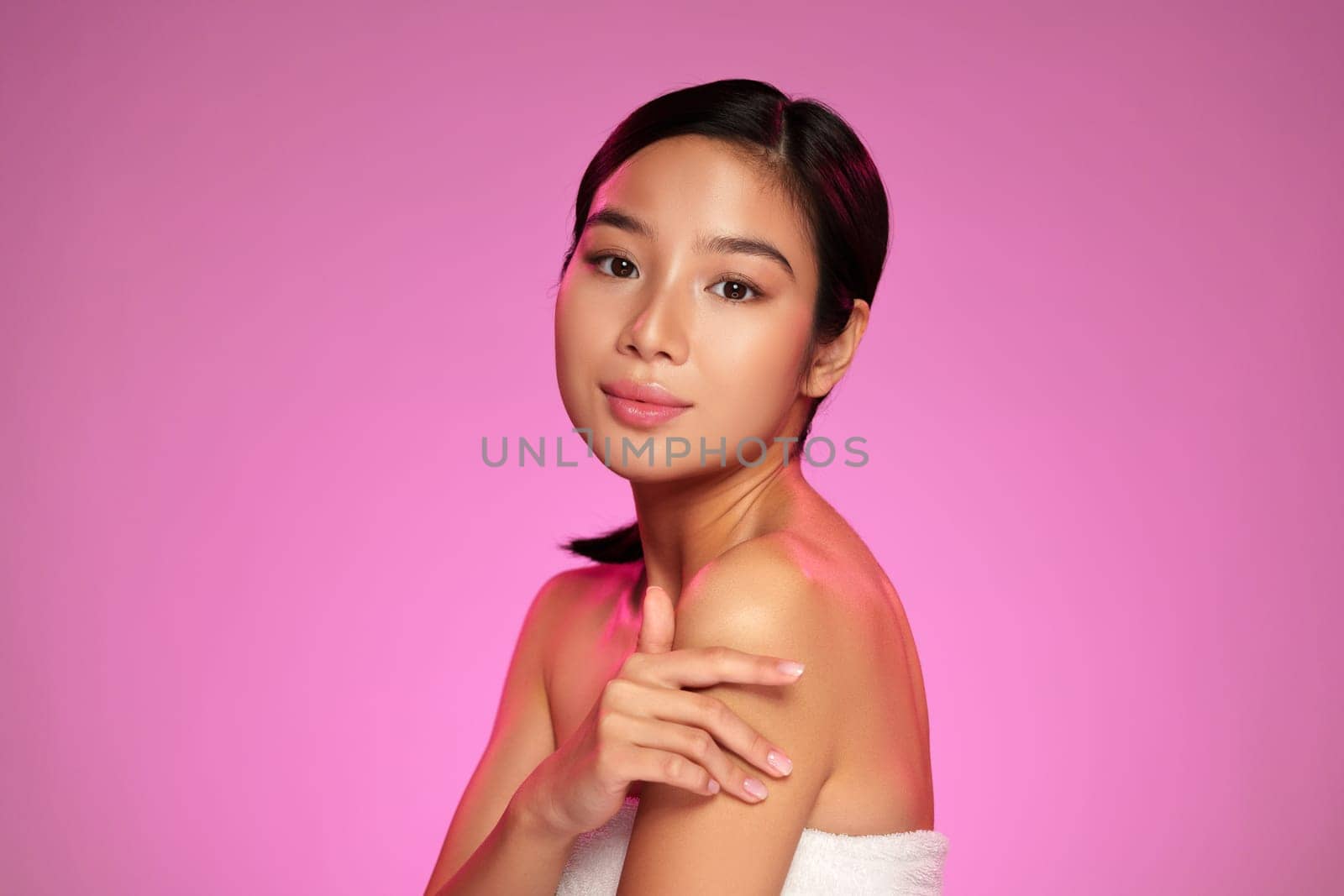 A young woman with smooth skin poses elegantly against a bright pink backdrop. She exudes confidence and tranquility while demonstrating a skincare routine with gentle gestures.