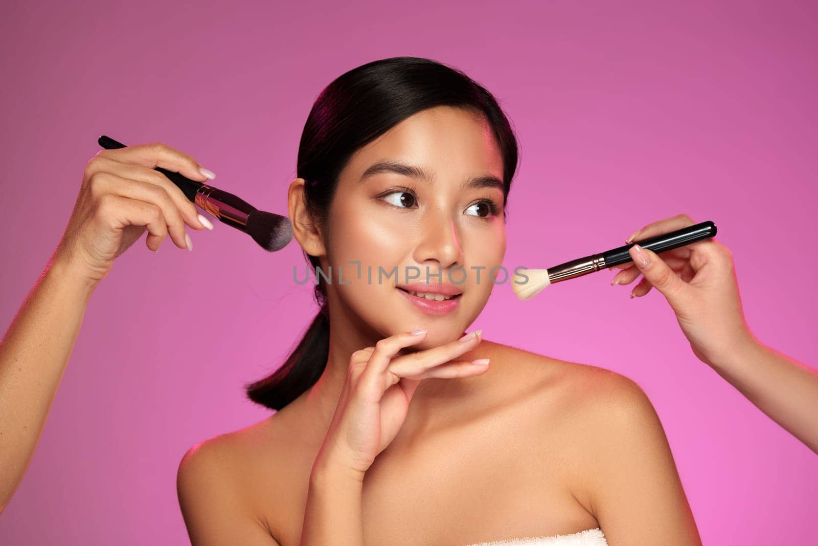 A model showcases a serene expression while receiving makeup application from two skilled artists. The backdrop is a bright pink, enhancing the beauty ritual atmosphere.