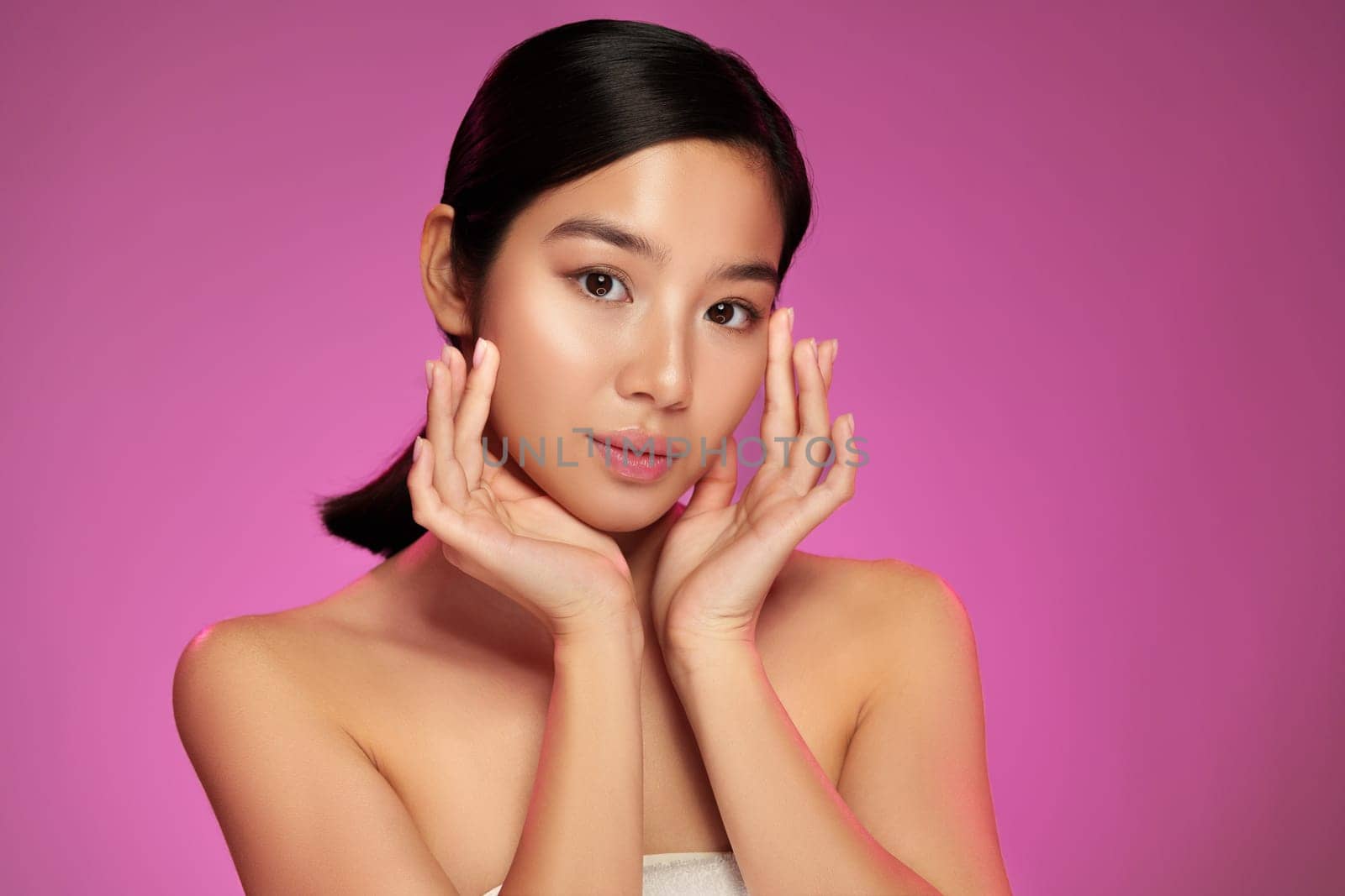 A young model poses elegantly, showcasing natural beauty with her hands gently framing her face. The vibrant pink background enhances her features, creating a striking visual contrast.