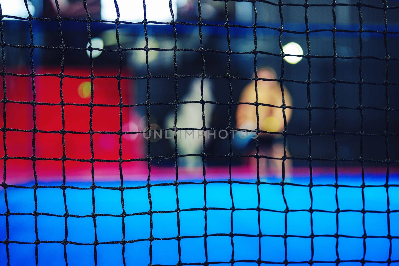 In a vibrant indoor sports facility, athletes train together against a backdrop of colorful surfaces. A net blurs the view of their activities, creating a dynamic atmosphere during the evening.