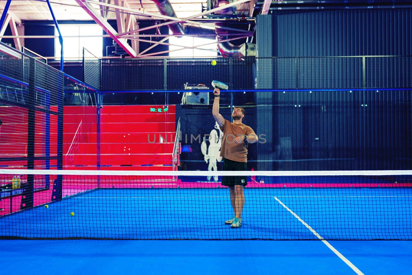 A male athlete prepares to serve a tennis ball in an indoor sports facility. The vibrant blue court contrasts with the red seating area in the background.