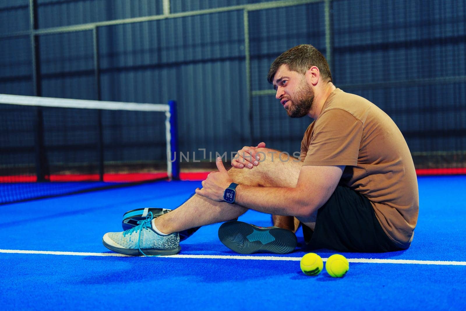 A man is seated on a vibrant blue tennis court, visibly in pain as he holds his ankle. Nearby, tennis balls lie on the ground, indicating a recent match.