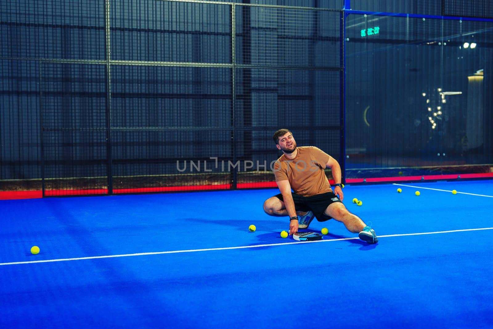 A man is engaged in an intense game of padel tennis, making a dynamic dive to reach a ball on a bright blue court. The atmosphere is lively and competitive as balls scatter around.