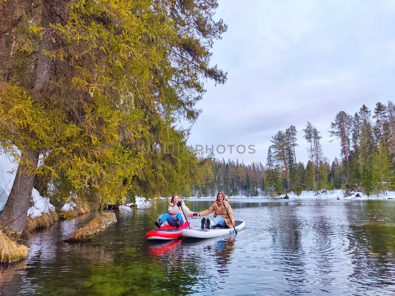 Kirov, Russia - March 14, 2024: A couple paddleboards together on a calm lake, surrounded by winter trees and a peaceful landscape under a cloudy sky.