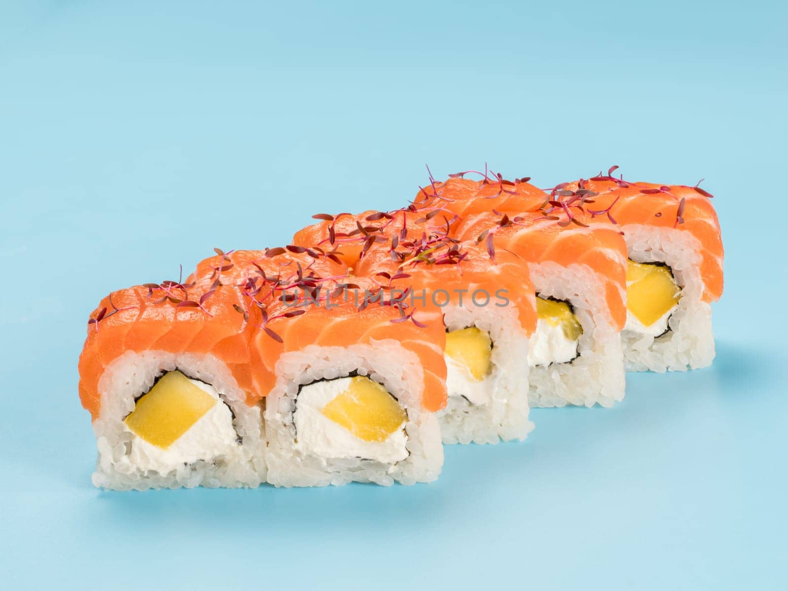 Studio photography of Philadelphia Roll of velvety cream cheese and mango wrapped in perfect seasoned sushi rice and fresh salmon, decorated microgreens. Isolated on solid light blue background