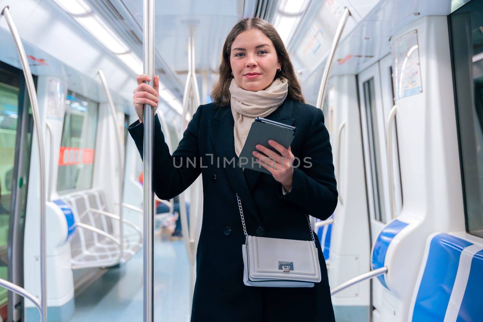 Stylish young woman in a black coat and beige scarf reading on an e-reader while standing inside a modern subway train. She balances while holding onto a pole during her commute.