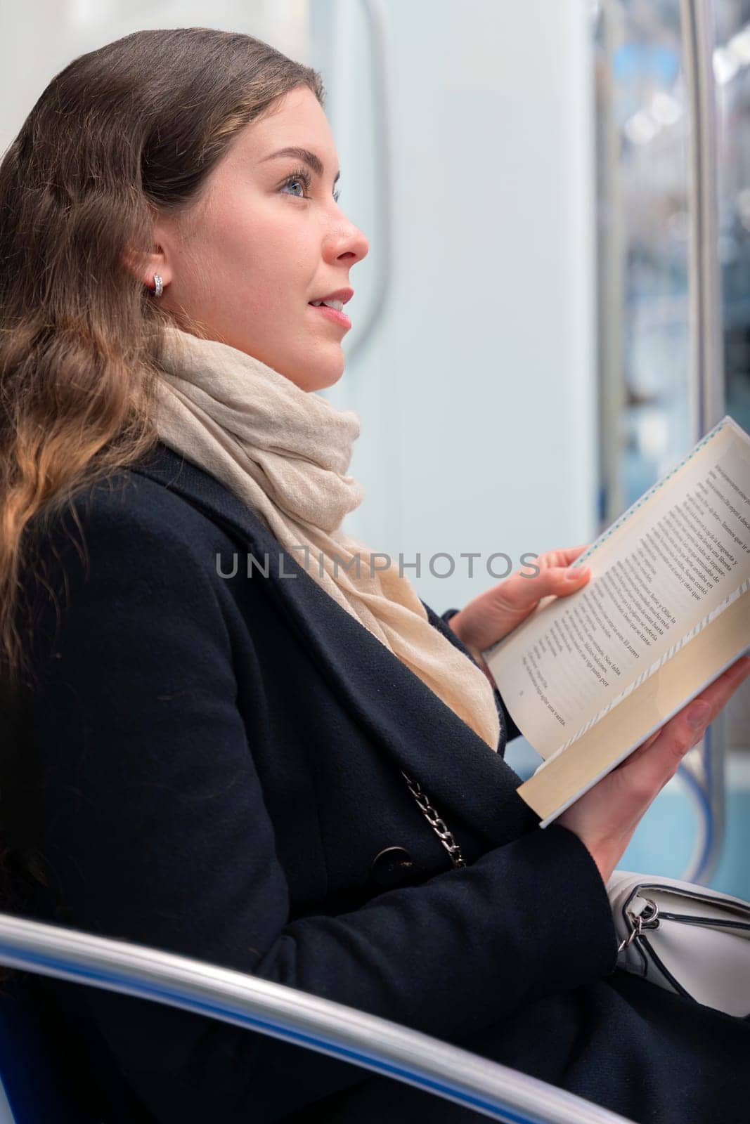 Young woman in a black coat and beige scarf sitting inside a modern subway train, reading a book with focus. She enjoys a quiet moment during her commute in the city.