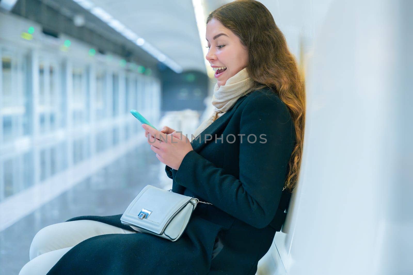 Stylish young woman in a black coat and scarf sitting in a modern subway station, smiling while using her smartphone. The bright and clean environment enhances the urban setting.