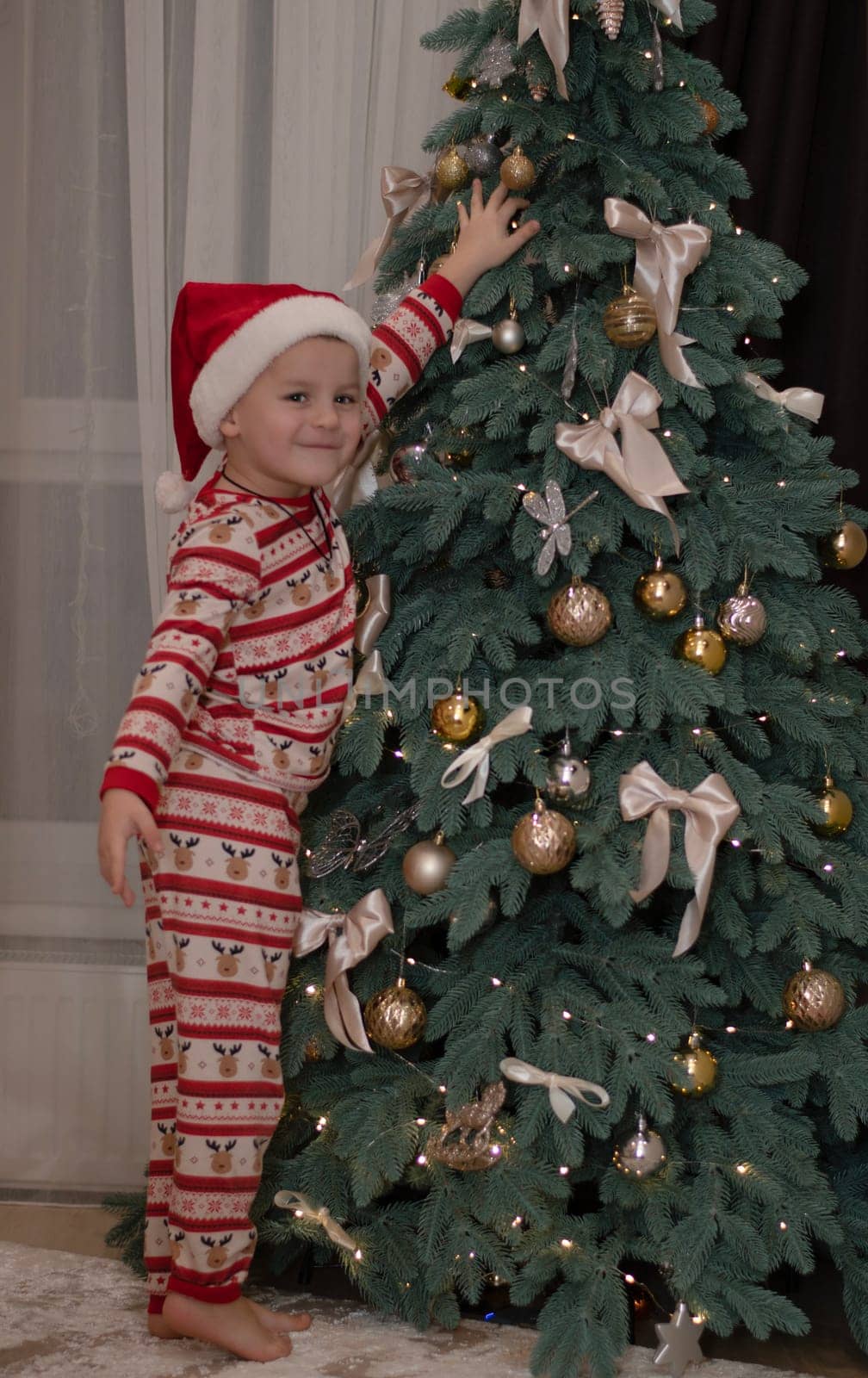Christmas concept. a 5-year-old boy in New Year's pajamas and a Santa Claus hat stands next to the Christmas tree and joyfully decorates it. Happy smile.