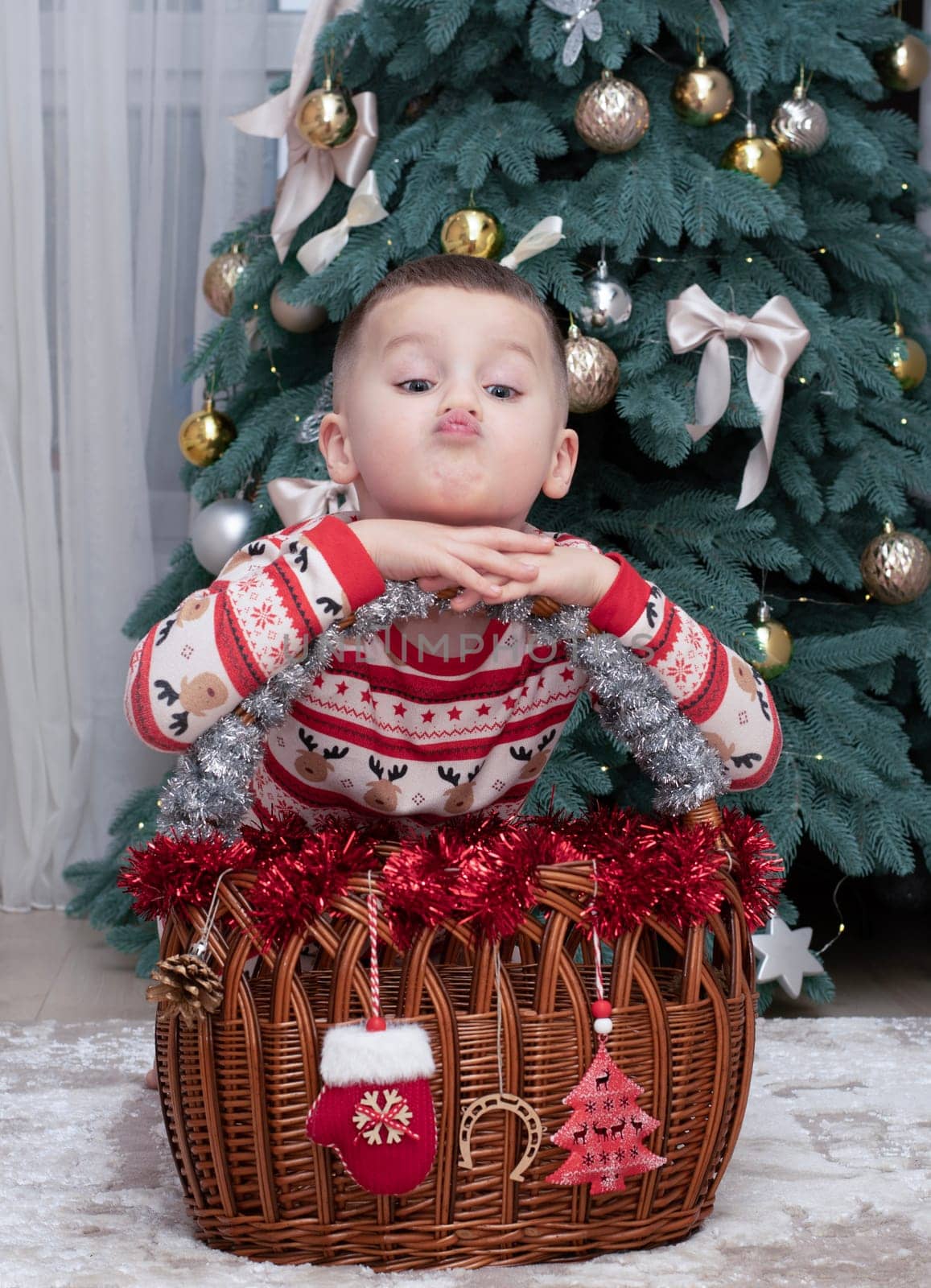 Christmas. A five-year-old boy in New Year's pajamas sits on the floor near a wicker New Year's basket against the backdrop of a Christmas tree and smiles cheerfully and makes faces. Facial expressions festive atmosphere