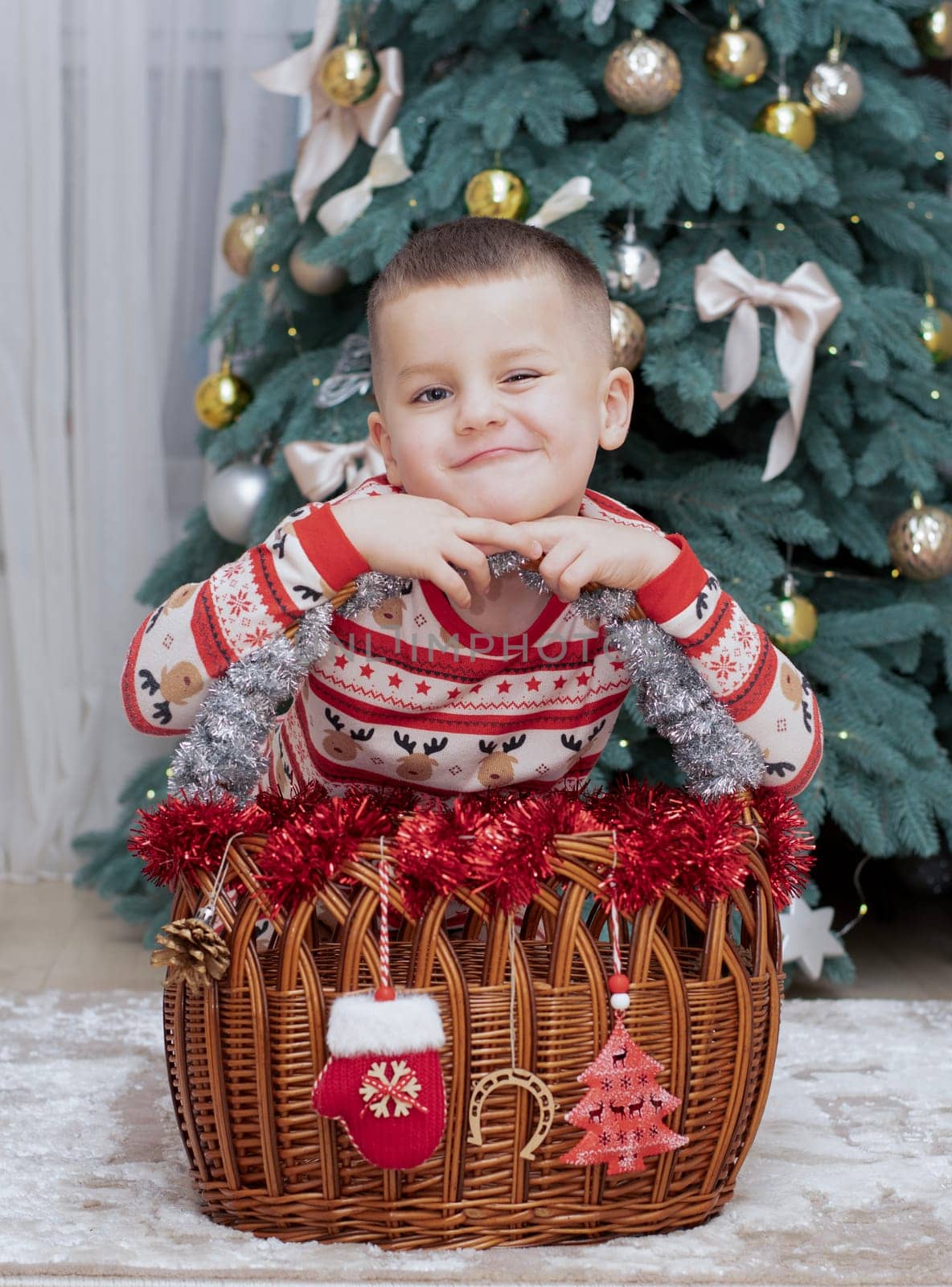 Christmas. A five-year-old boy in New Year's pajamas sits on the floor near a wicker New Year's basket against the backdrop of a Christmas tree and smiles cheerfully and makes faces. Facial expressions festive atmosphere