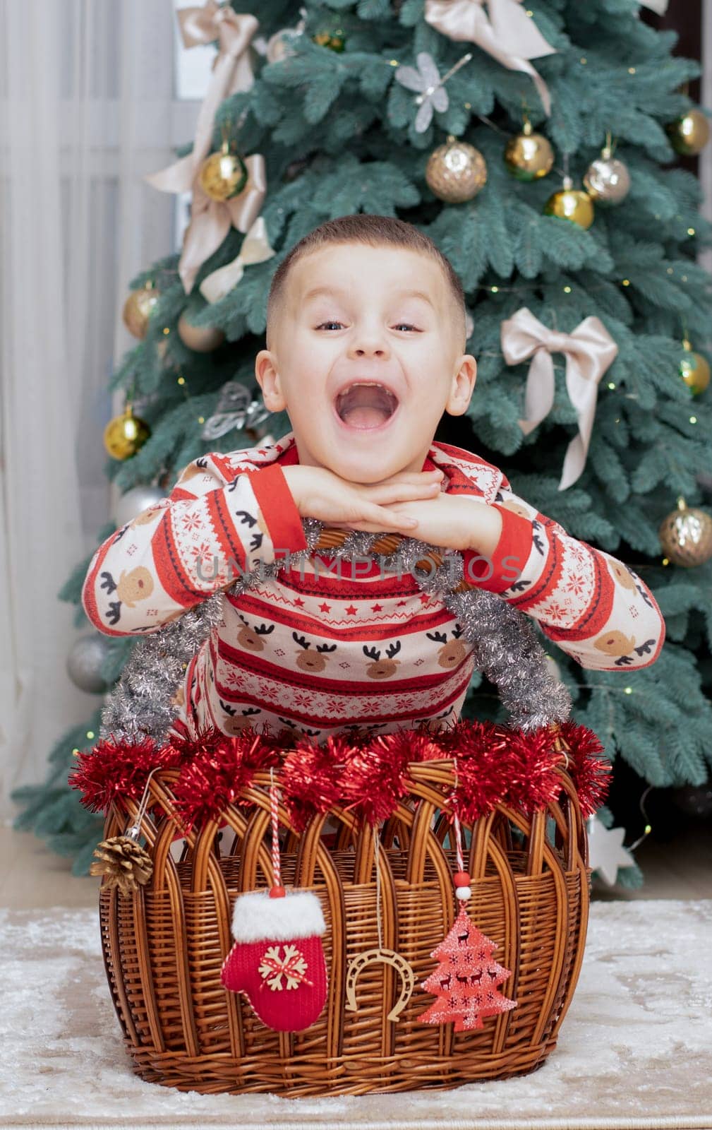 Christmas. A five-year-old boy in New Year's pajamas sits on the floor near a wicker New Year's basket against the backdrop of a Christmas tree and smiles cheerfully and makes faces. Facial expressions festive atmosphere