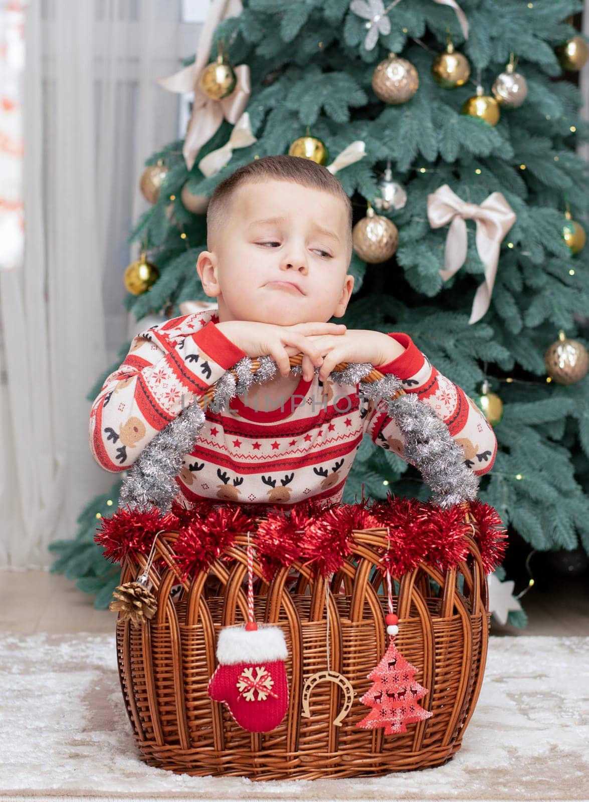 Christmas. A five-year-old boy in New Year's pajamas sits on the floor near a wicker New Year's basket against the backdrop of a Christmas tree and smiles cheerfully and makes faces. Facial expressions festive atmosphere