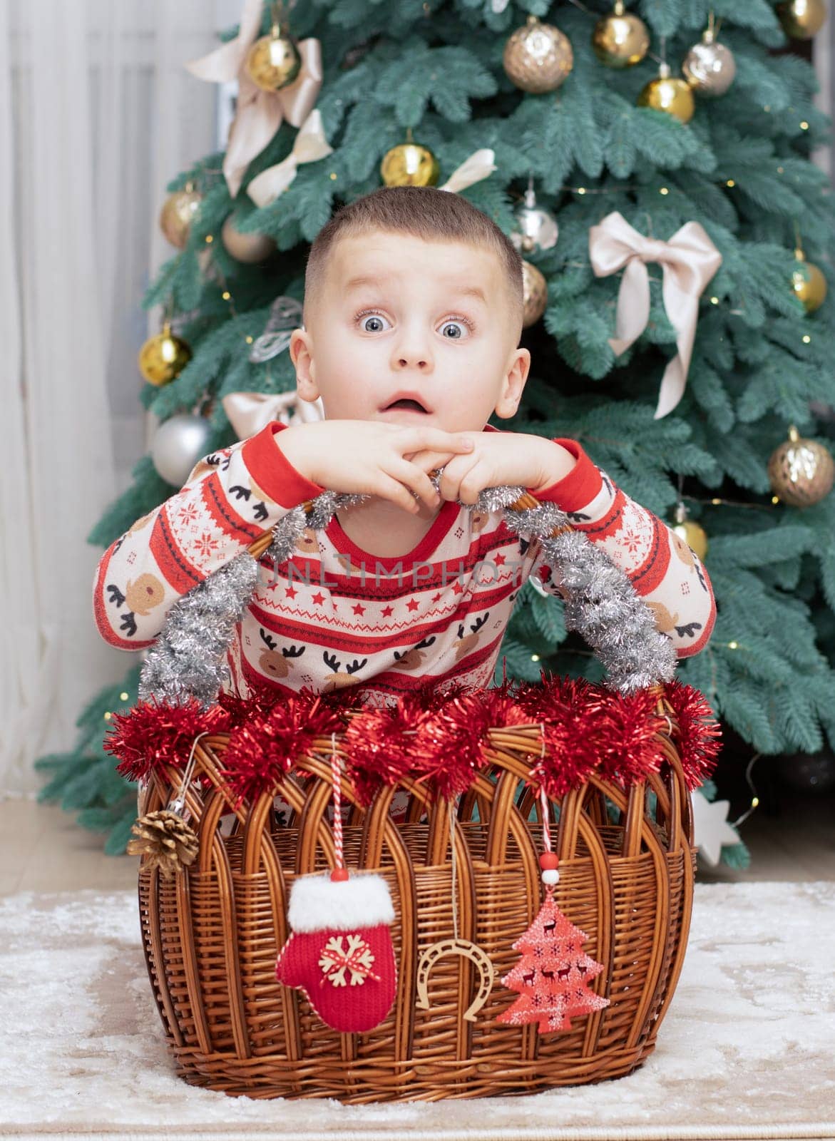 Christmas. A five-year-old boy in New Year's pajamas sits on the floor near a wicker New Year's basket against the backdrop of a Christmas tree and smiles cheerfully and makes faces. Facial expressions festive atmosphere