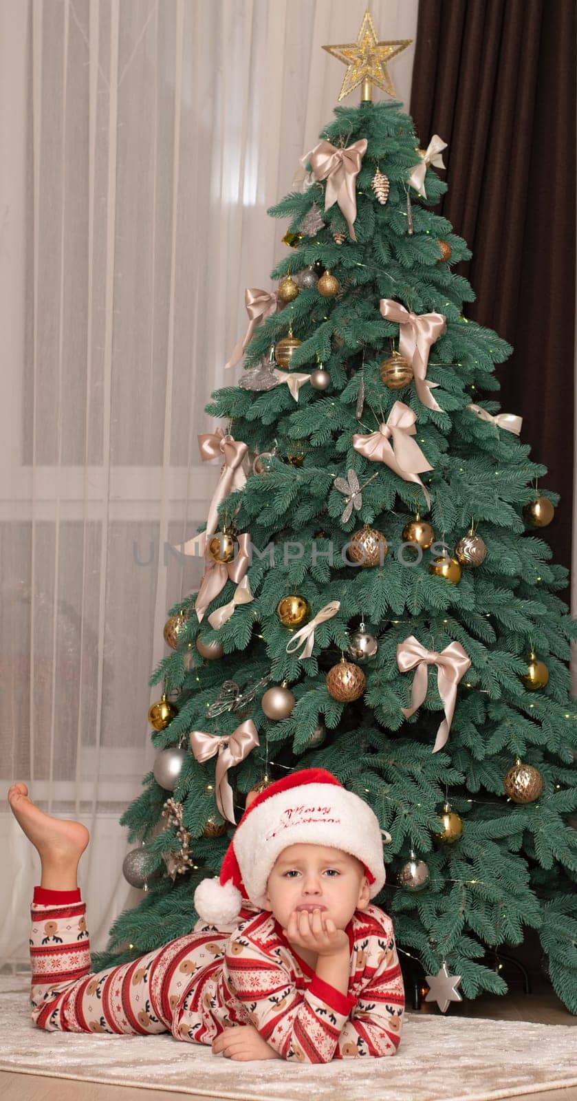 Christmas. A five-year-old boy in New Year's pajamas lies on the floor near the Christmas tree in a Santa Claus hat and smiles cheerfully. Facial expressions of joy and happiness. festive atmosphere