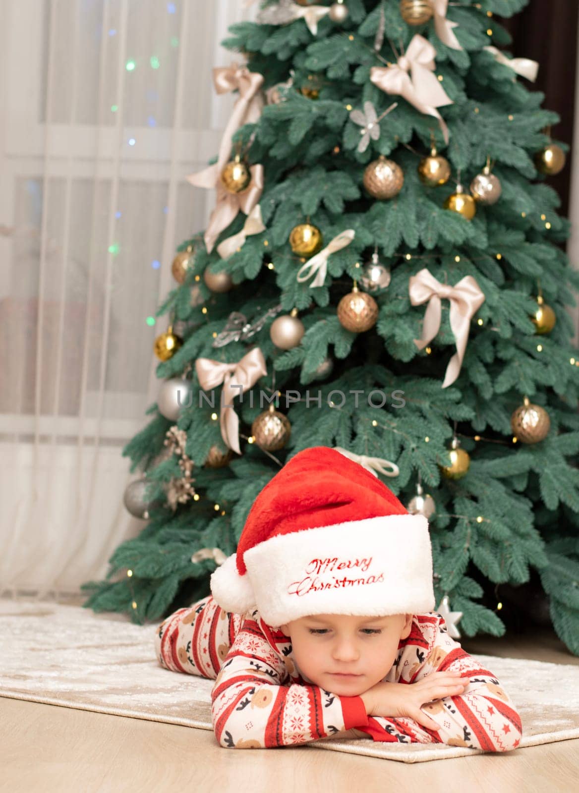 Christmas. A five-year-old boy in New Year's pajamas lies on the floor near the Christmas tree wearing a Santa Claus hat. Pensive, sad, dreamy. festive atmosphere