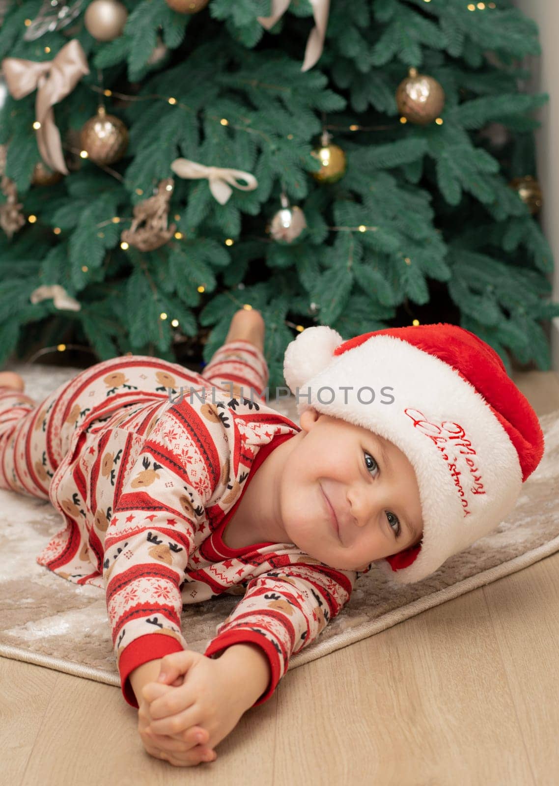 Christmas. A five-year-old boy in New Year's pajamas lies on the floor near the Christmas tree in a Santa Claus hat and smiles cheerfully. Facial expressions of joy and happiness. festive atmosphere