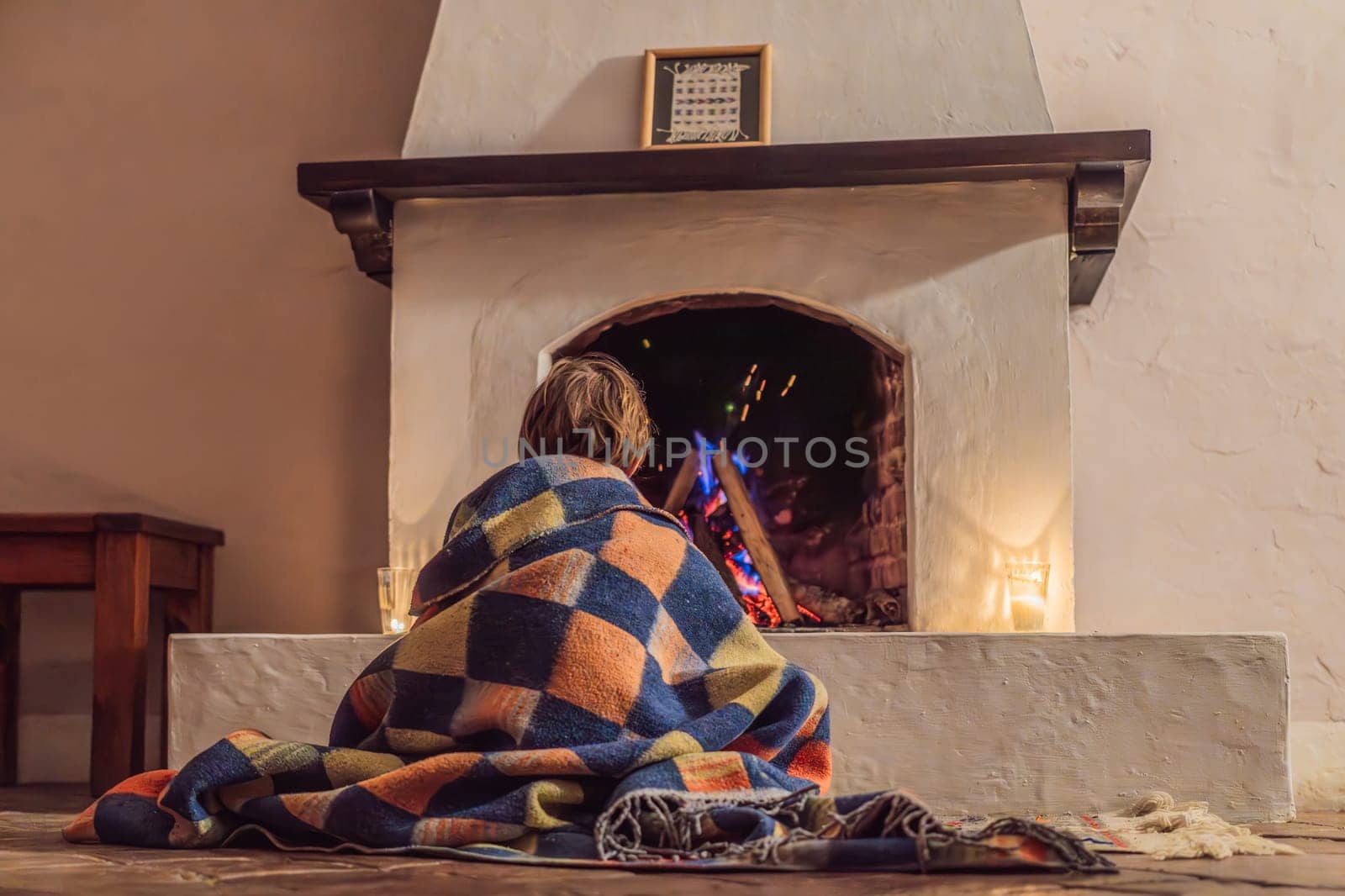 Boy by the fireplace during cozy winter evening. Warm and peaceful family moment indoors. Togetherness, love, and relaxation concept. A warm and inviting scene featuring a lit fireplace with glowing firewood and candles. A colorful traditional rug and pillow lie on the floor, creating a cozy atmosphere. Home, relaxation, and rustic comfort concept.