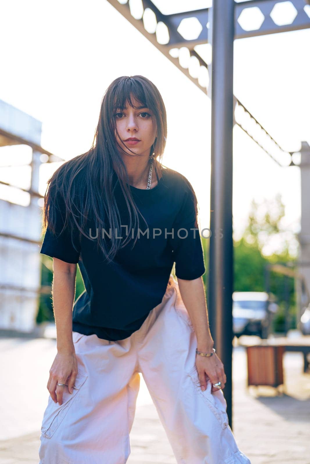 A young woman stands outdoors in an urban area, showcasing a casual outfit with a relaxed posture. The sun sets in the background, creating a warm ambiance.