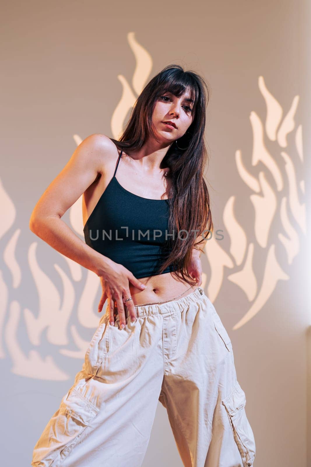 A woman with long hair poses confidently in a stylish tank top and loose cargo pants, surrounded by artistic flame shadows on the wall. She exudes energy and presence.