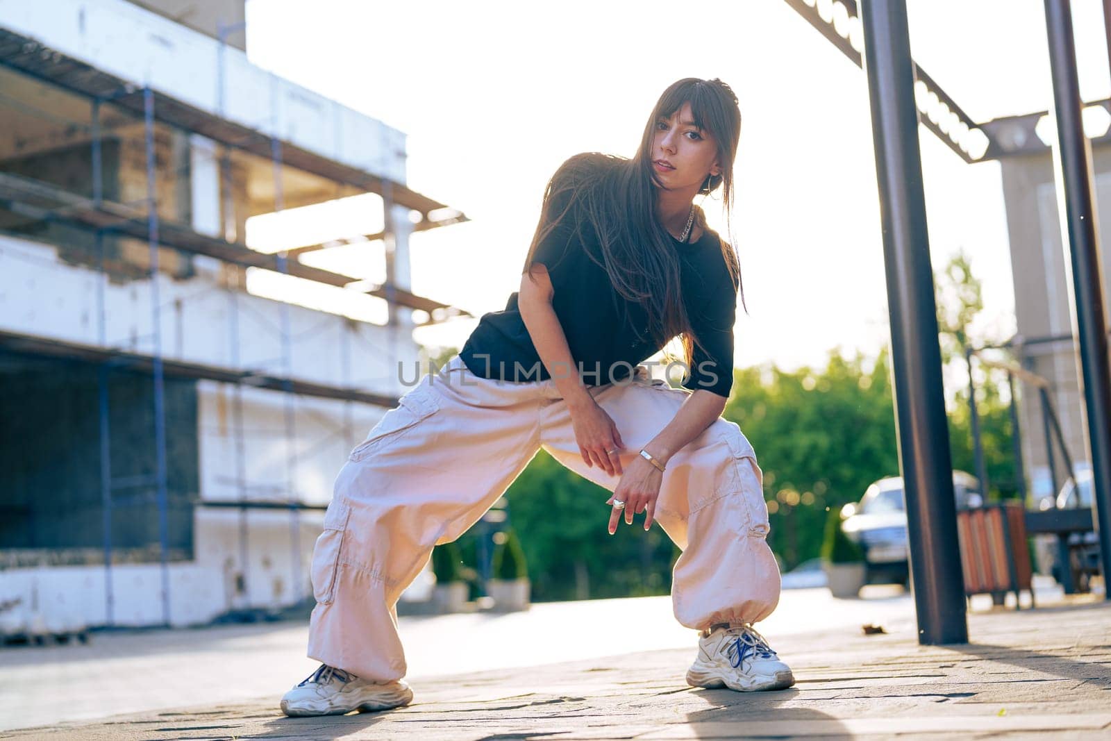A young woman showcases her dance skills in baggy pants and a black top at a construction site illuminated by the warm glow of sunset. Her confident pose captures the energy of urban dance.