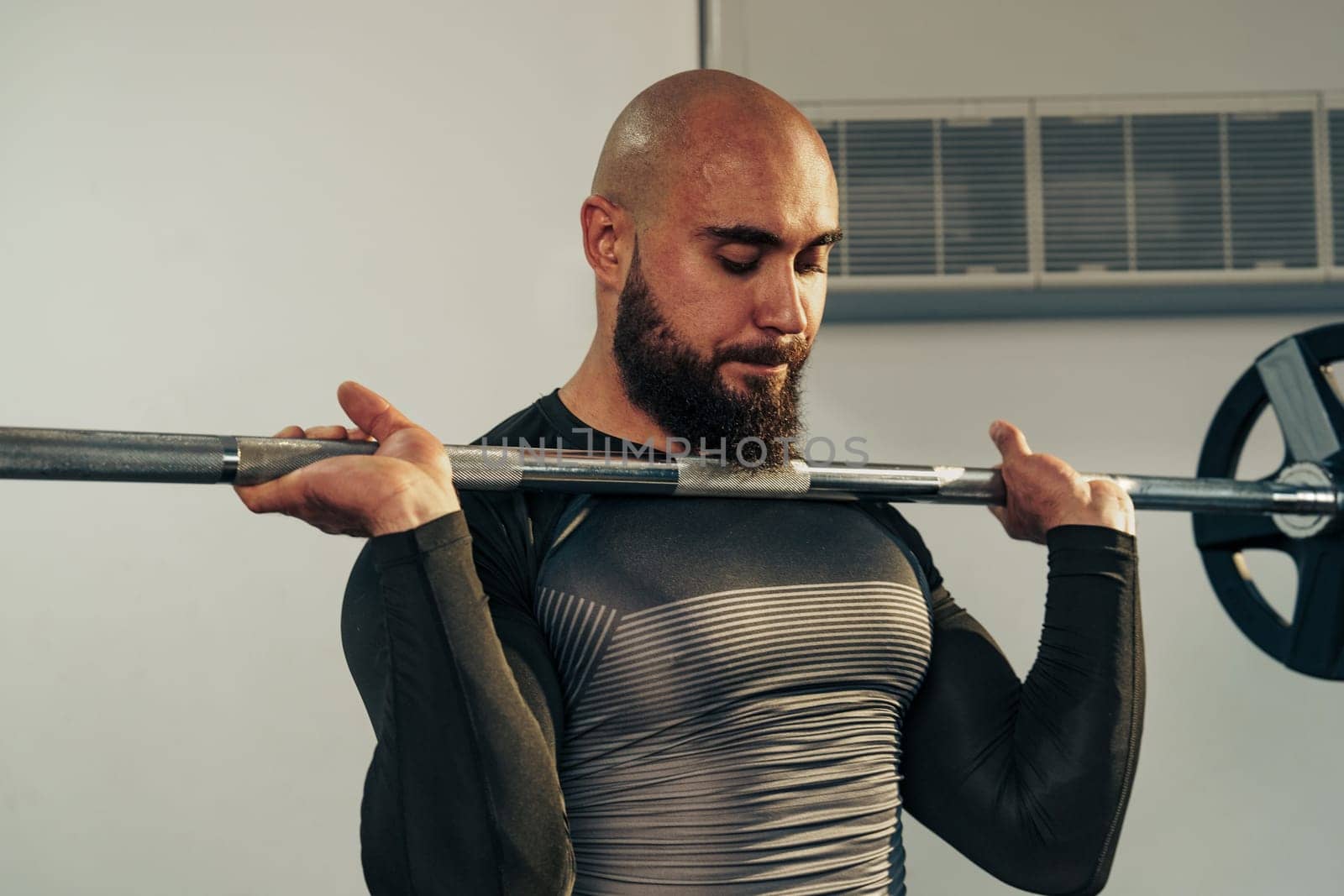 A man with a beard is performing a bicep curl with a barbell in a gym. He is focused and wearing a fitted shirt, showcasing his athletic physique during an intense workout.