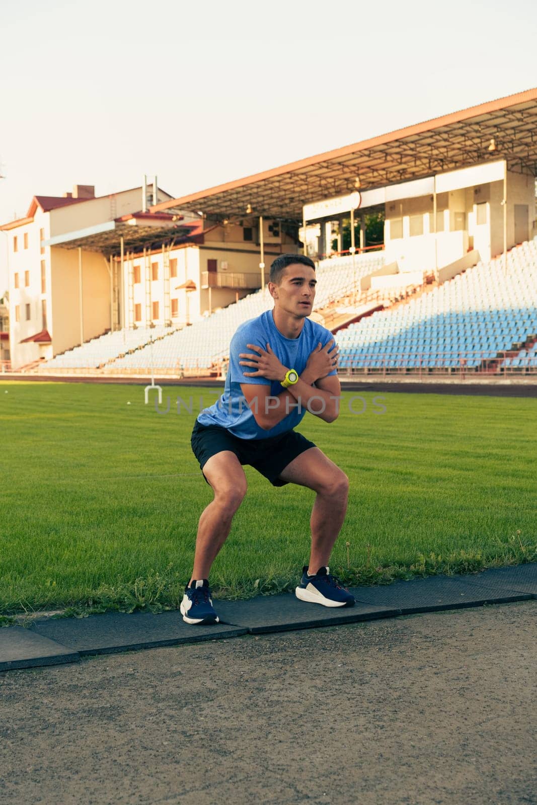 A man in a blue shirt and black shorts is engaged in a squat exercise on a track field. The sun sets, creating a warm glow over the empty stadium seating.