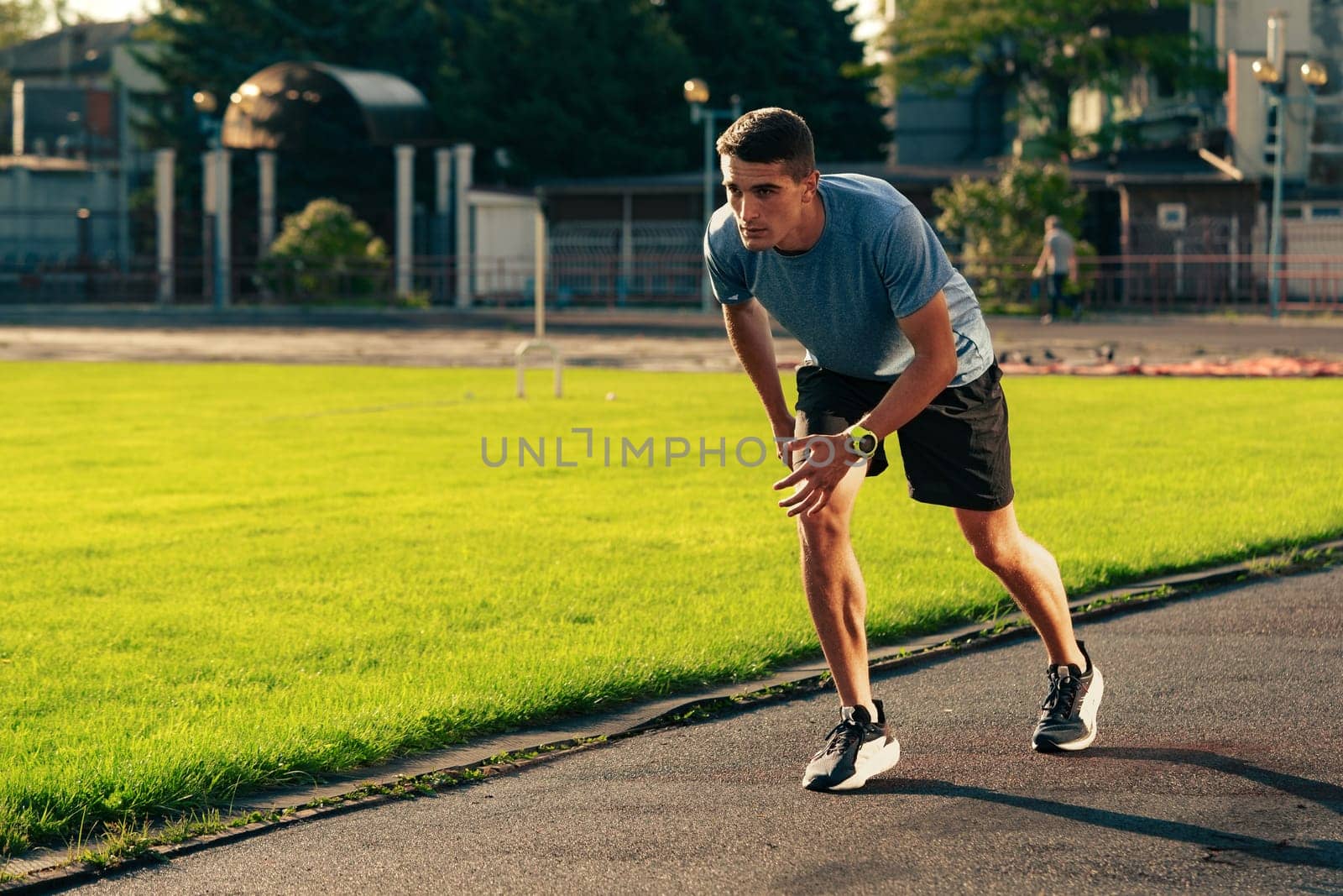 A man is sprinting on a running track located in an outdoor athletic facility. The sun is setting, casting warm light over the grass and track, creating an energetic atmosphere.