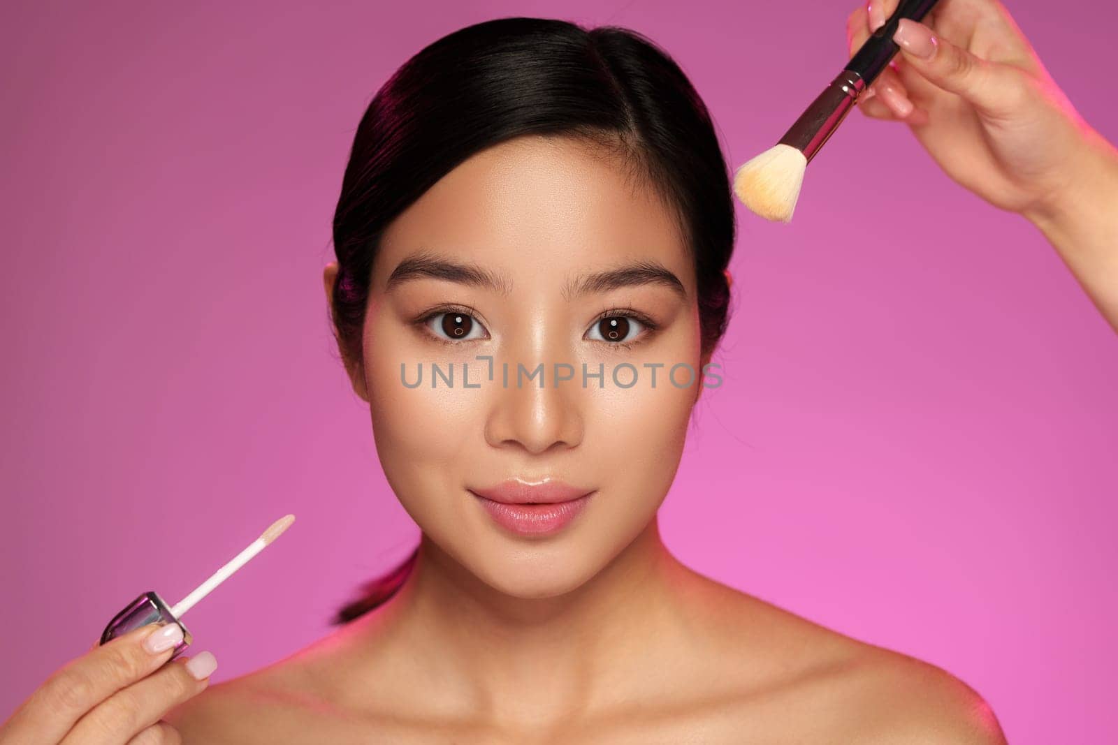 A model is having her makeup applied with glowing skin. One hand holds a brush while another showcases a cosmetic product. The backdrop features a vibrant pink hue.