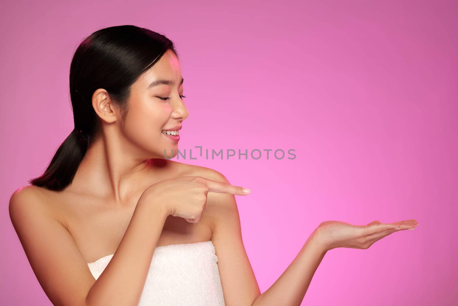 A young woman poses confidently with a warm smile, pointing at her palm. The soft pink background enhances the playful and inviting atmosphere.