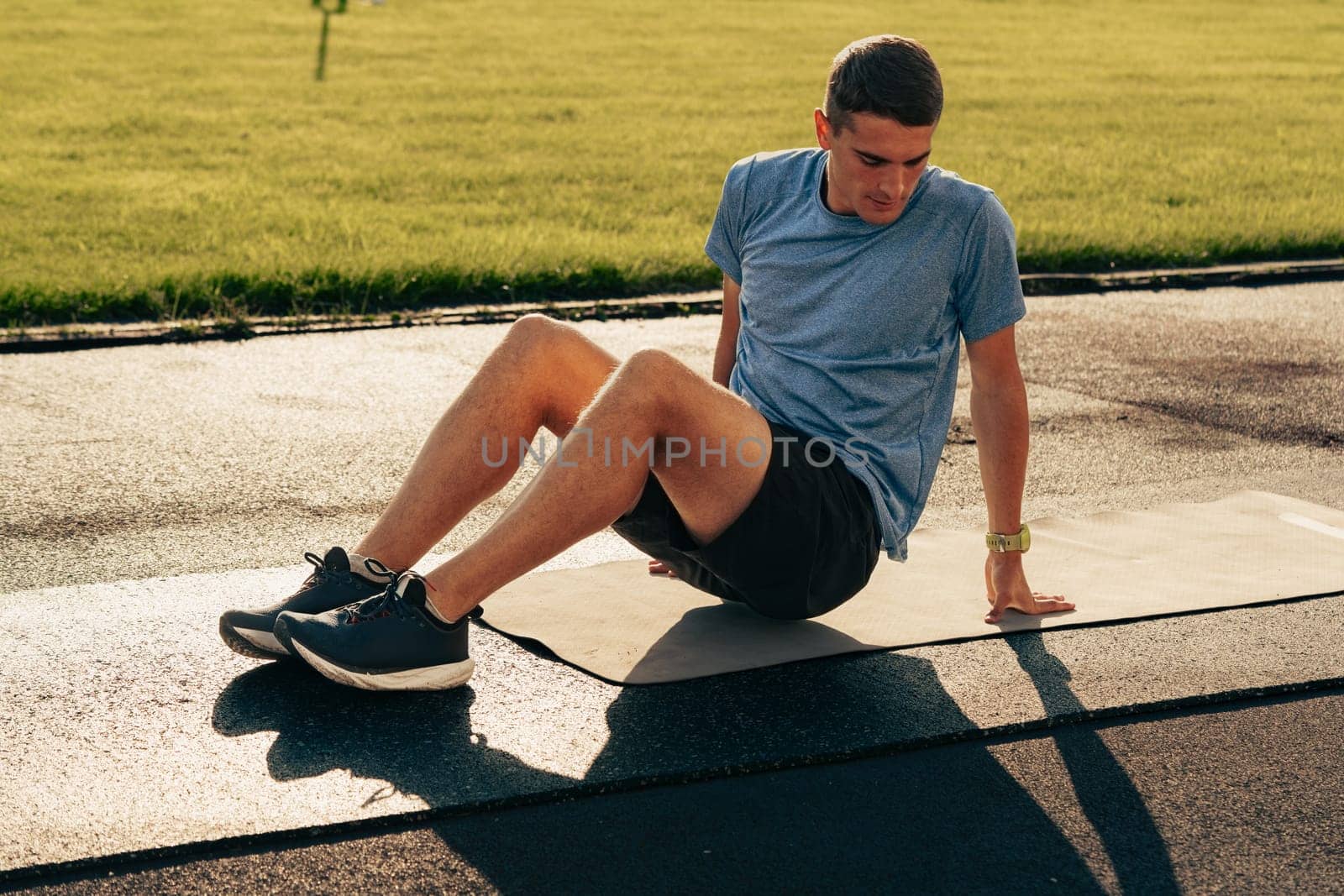 A young man is engaged in fitness training, performing exercises on a mat in a park. The sunny afternoon creates a vibrant atmosphere while he focuses on building strength.