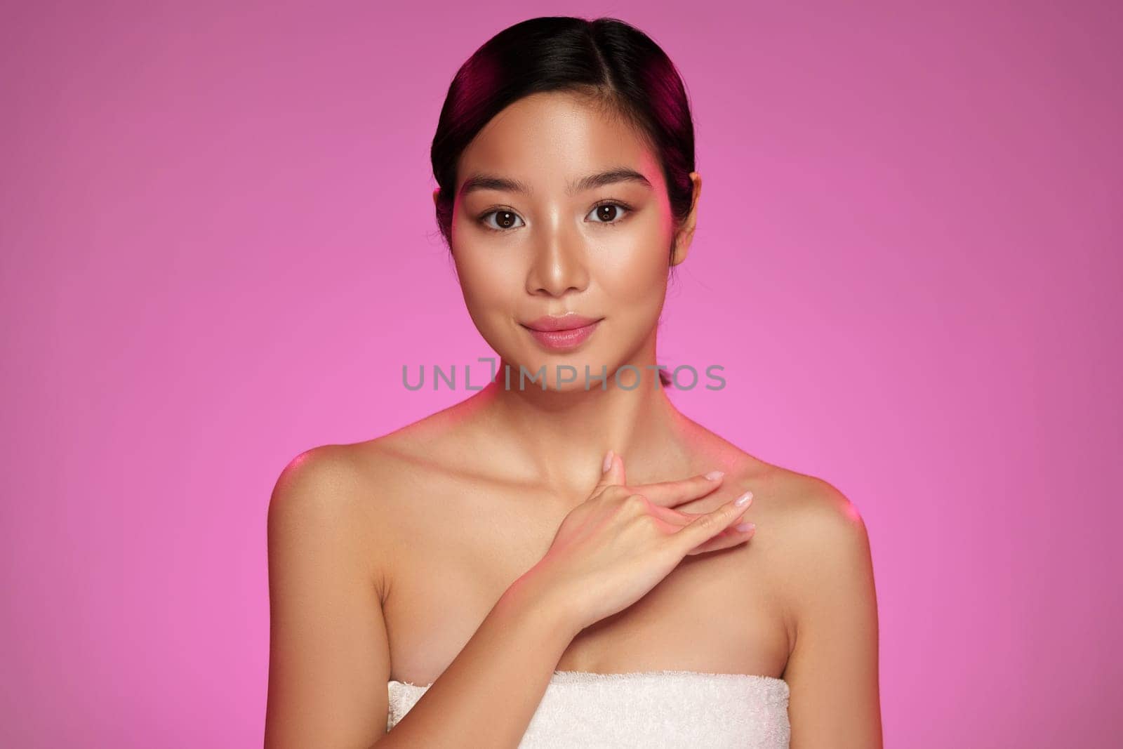 A young woman stands confidently with her hand near her neck against a soft pink backdrop. Her glowing skin and serene expression highlight her beauty in a minimalist setting.