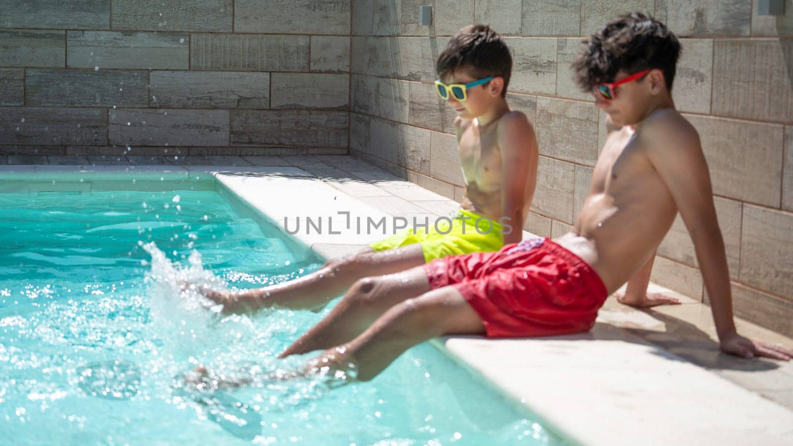 Two young boys wearing colorful swim trunks and sunglasses are relaxing by the pool, splashing their feet in the refreshing water on a sunny summer day.