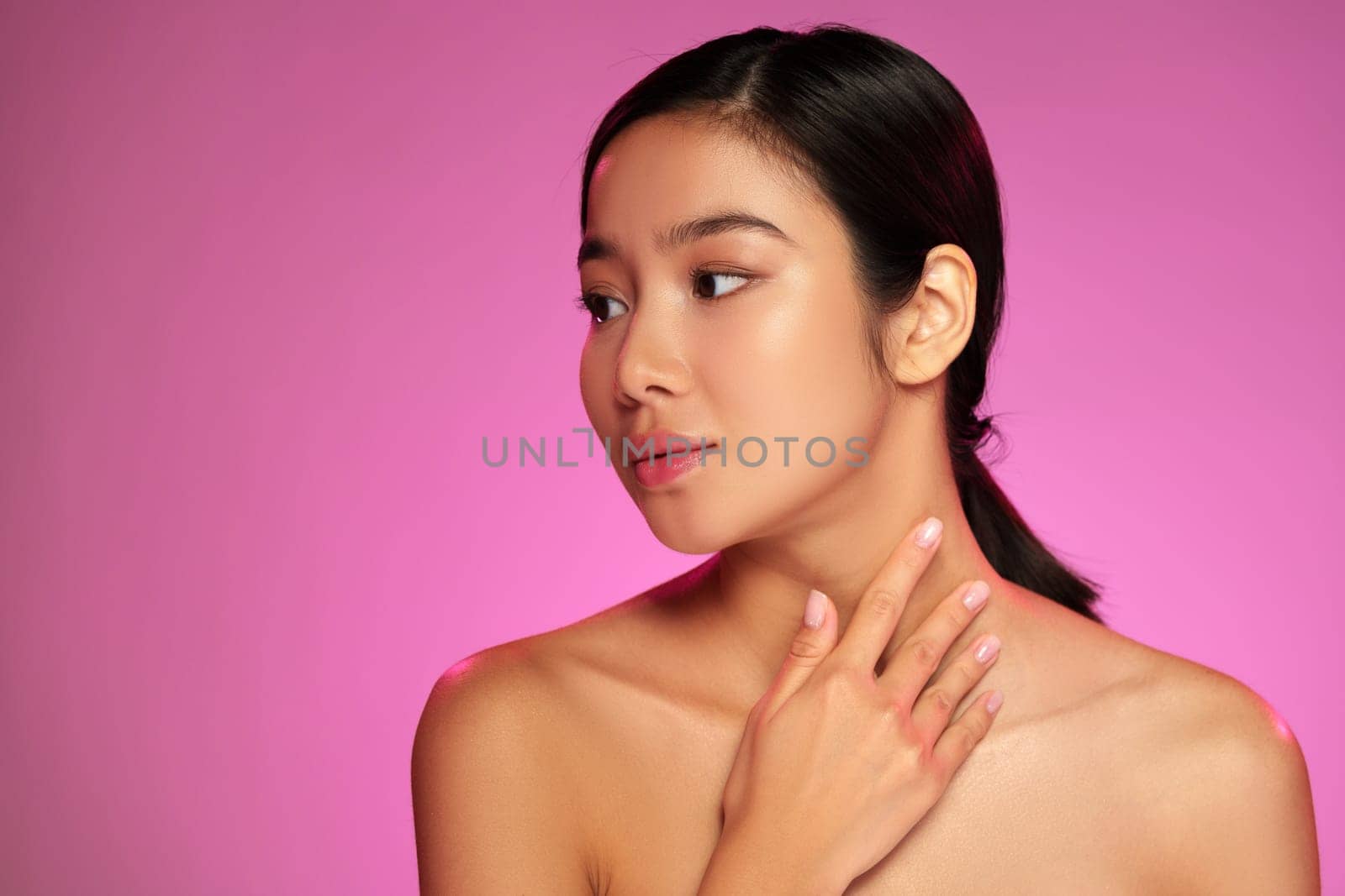 A young woman with smooth skin poses delicately, her hand resting on her neck. The background is a striking pink, highlighting her features and creating a captivating atmosphere.