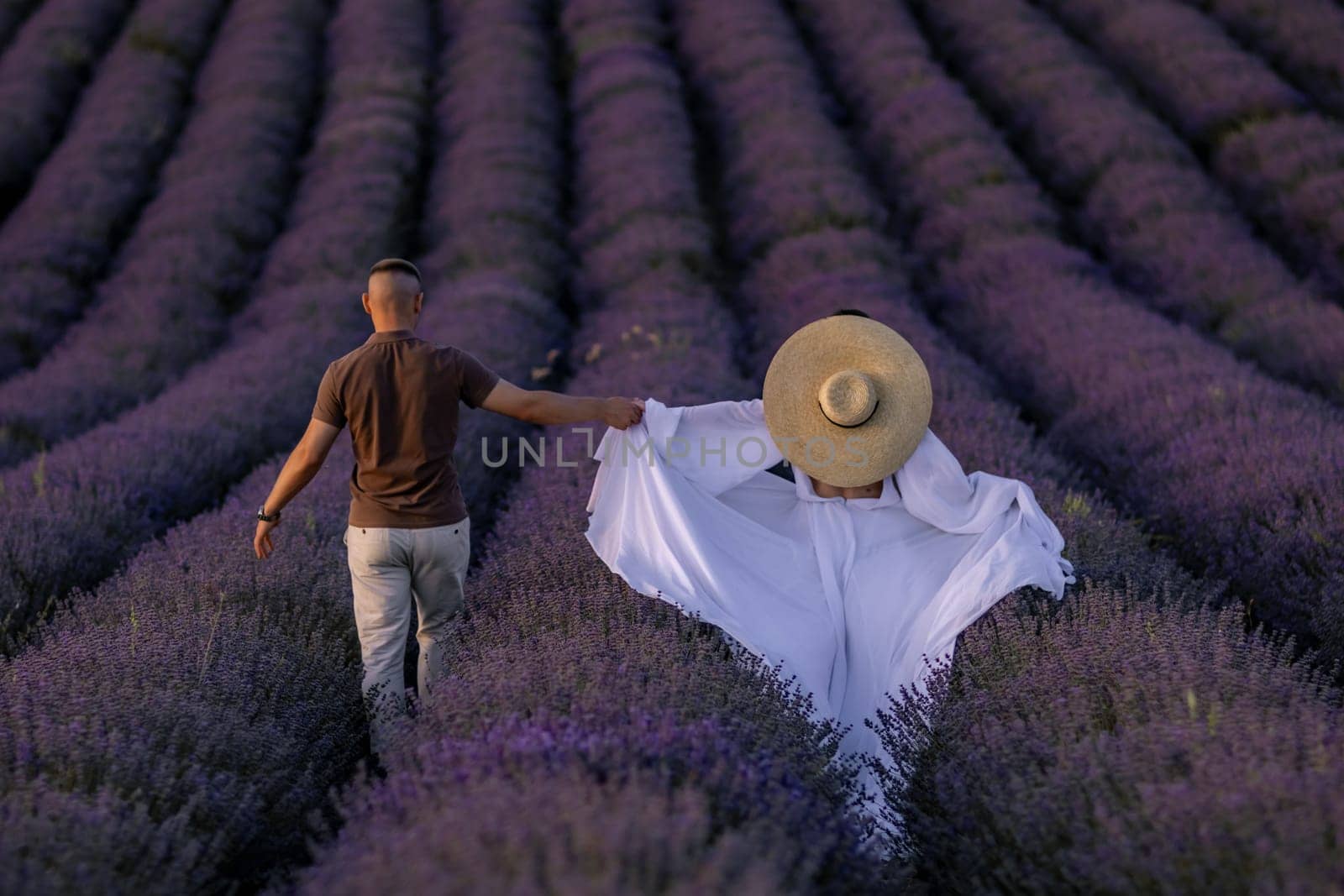 Here's a caption describing the image in under 150 characters: Man and woman stroll hand-in-hand through a vast, vibrant lavender field. She wears a wide-brimmed hat and flowing white fabric.