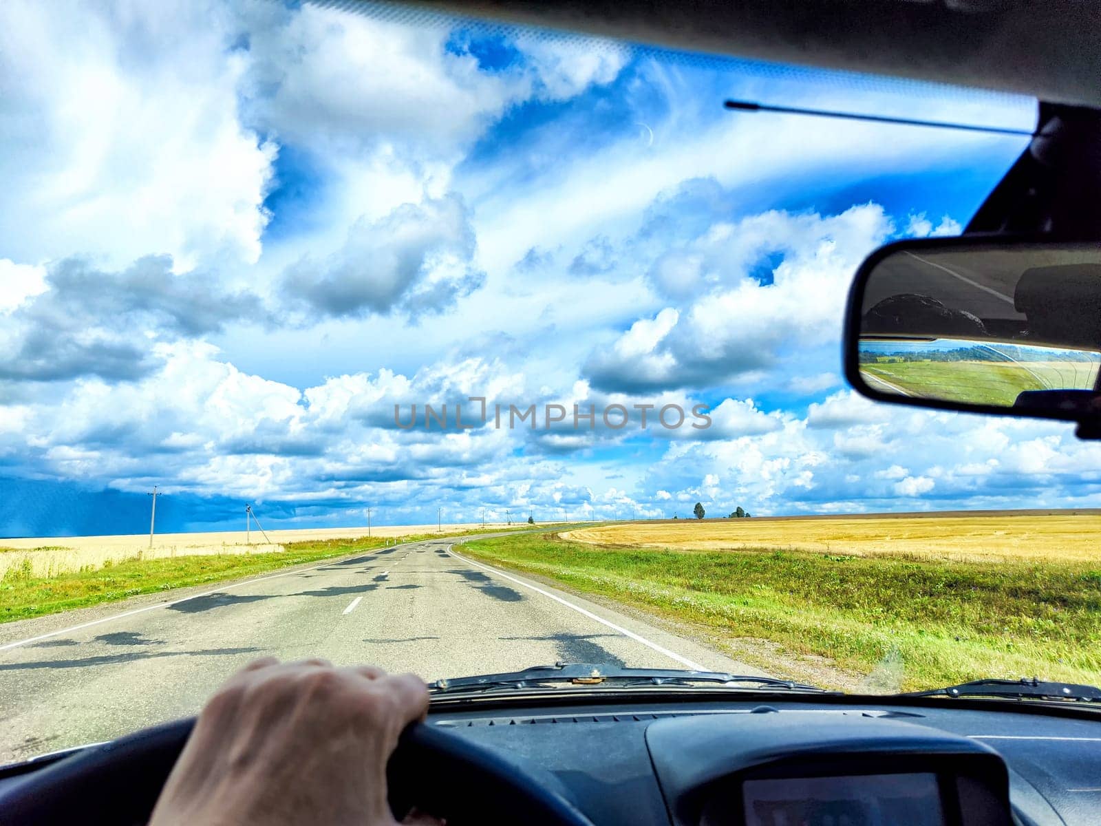 A hand grips the steering wheel while driving along wide road surrounded by fields under a beautiful blue sky