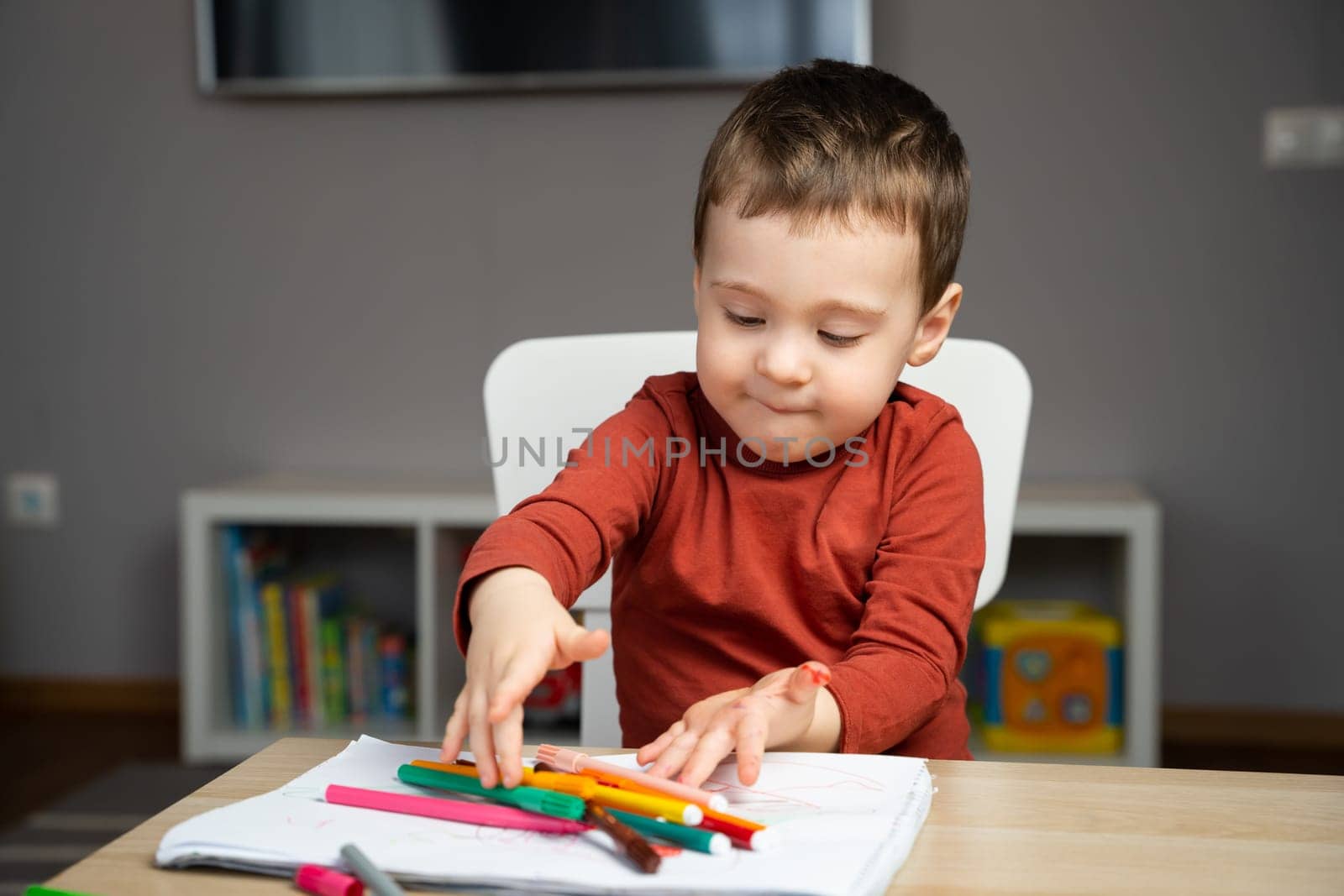 A cute happy little toddler boy of two years playing with markers in the album in the children's room at home. Educational activities for kids. Selective focus.
