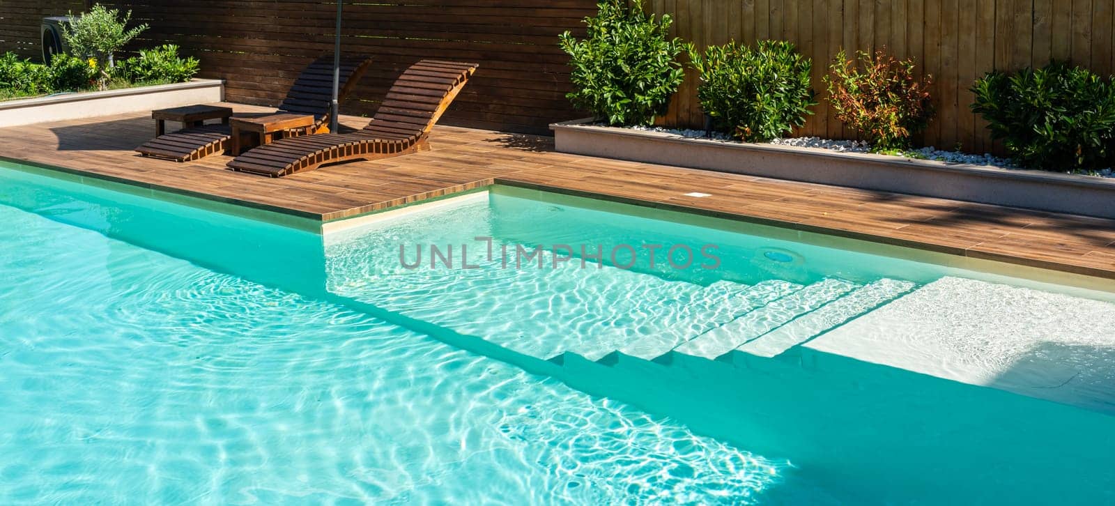 Modern swimming pool with turquoise water, wooden deck, and relaxing lounge chairs under sunny sky