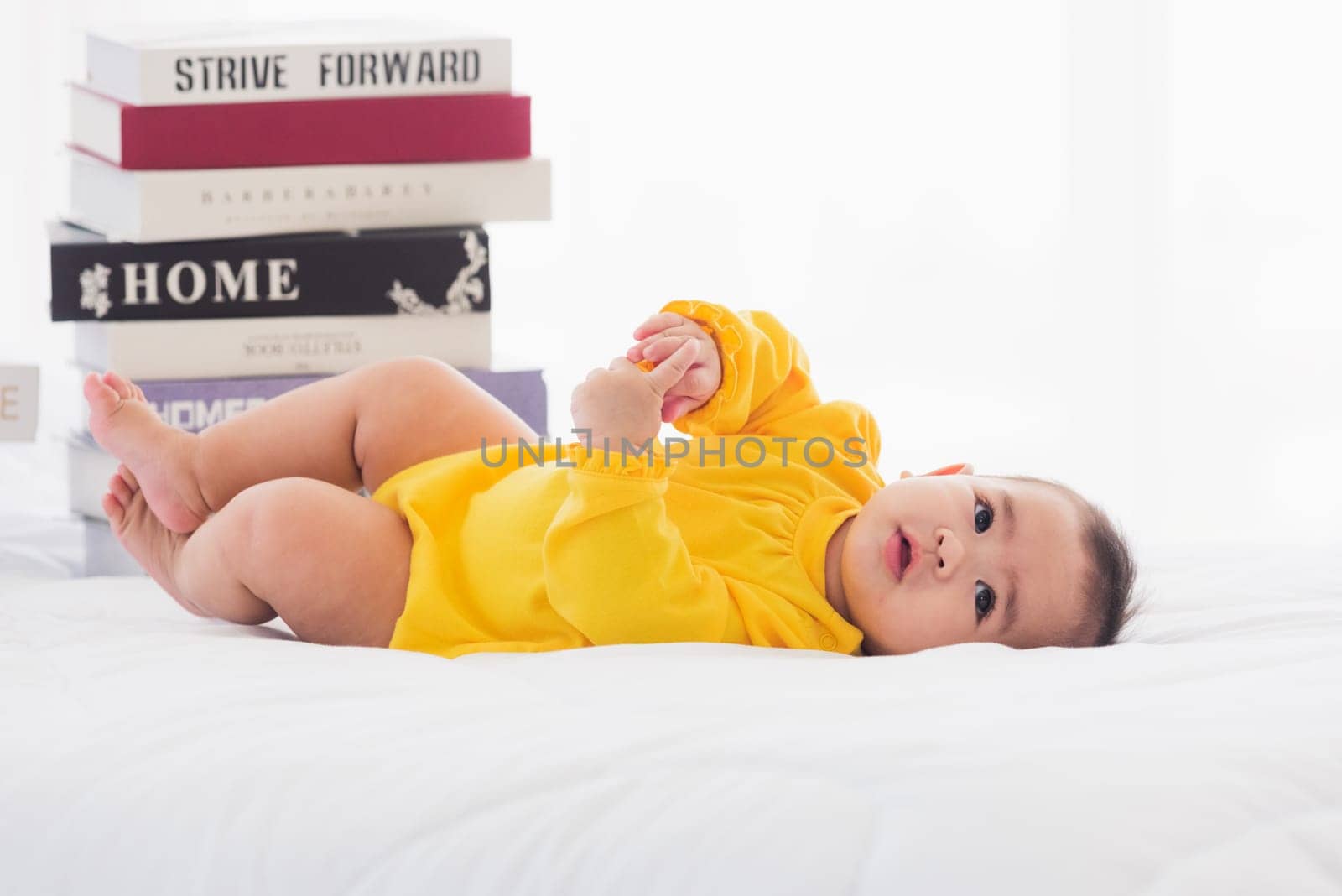 Asian cute baby little girl toddler looking at a stack of books on bedroom white bed, First baby library concept