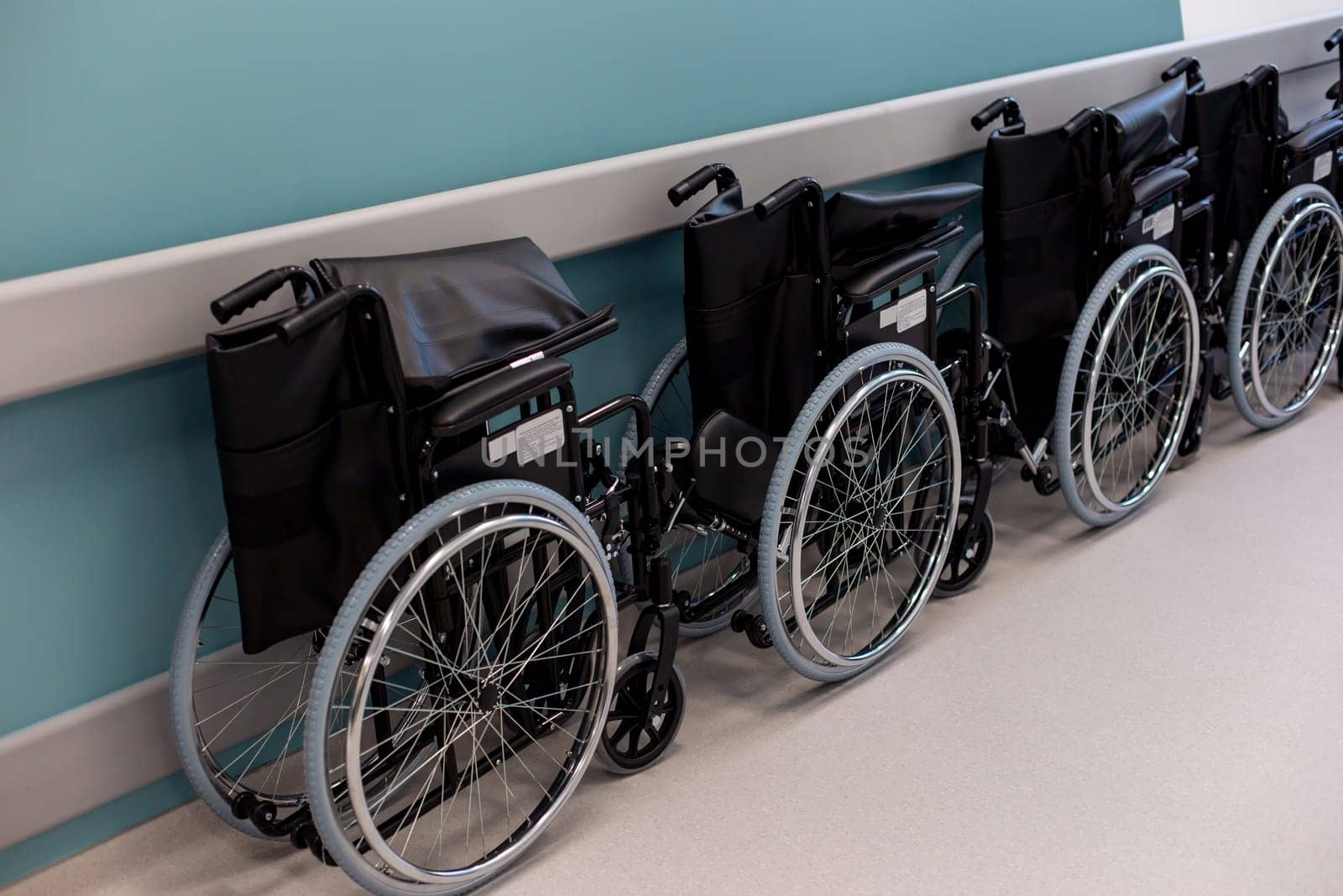 A row of empty folded black wheelchairs along a corridor with a blue wall.