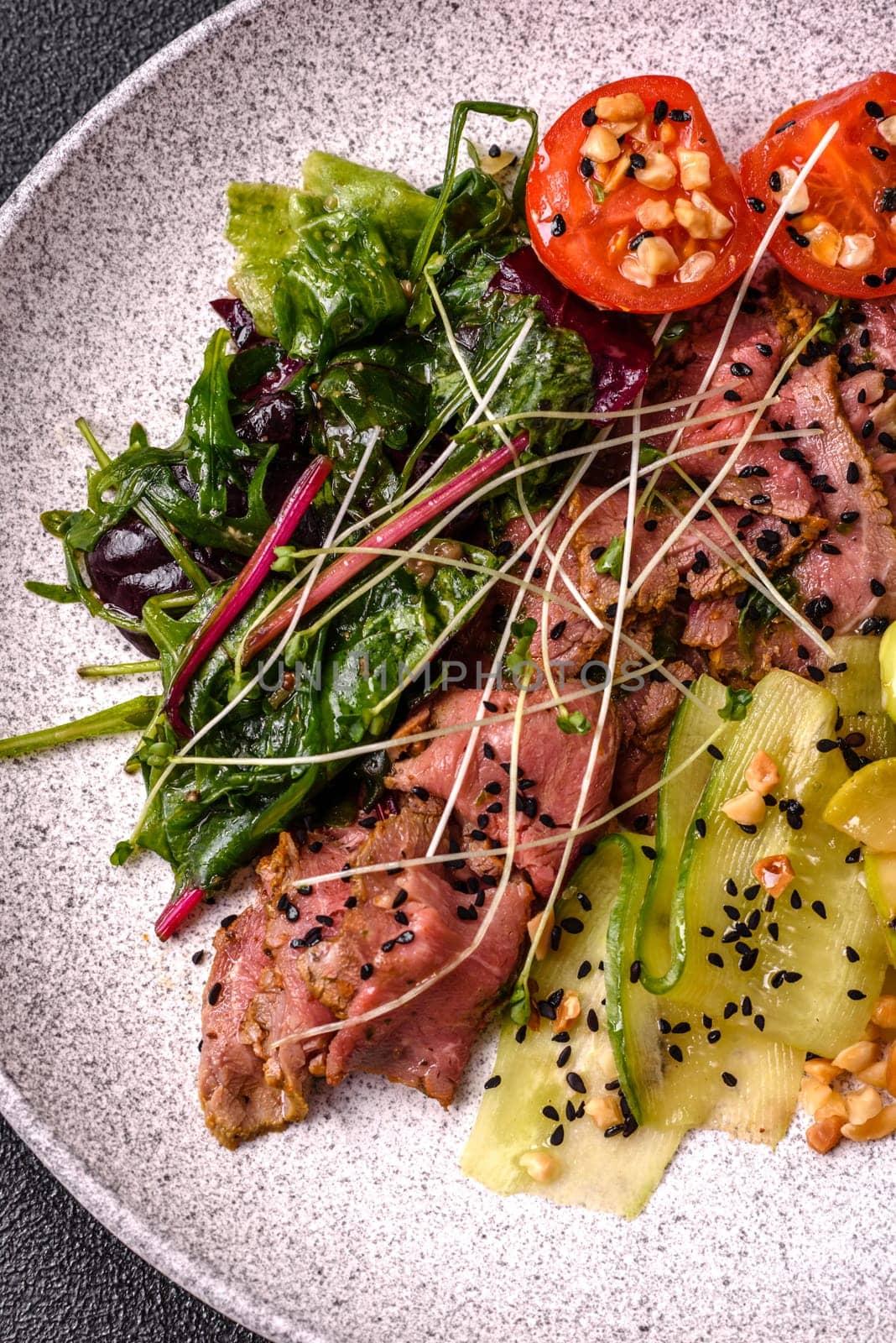 Warm salad consisting of slices of roast beef, greens, tomatoes, cucumbers, green beans on a dark concrete background