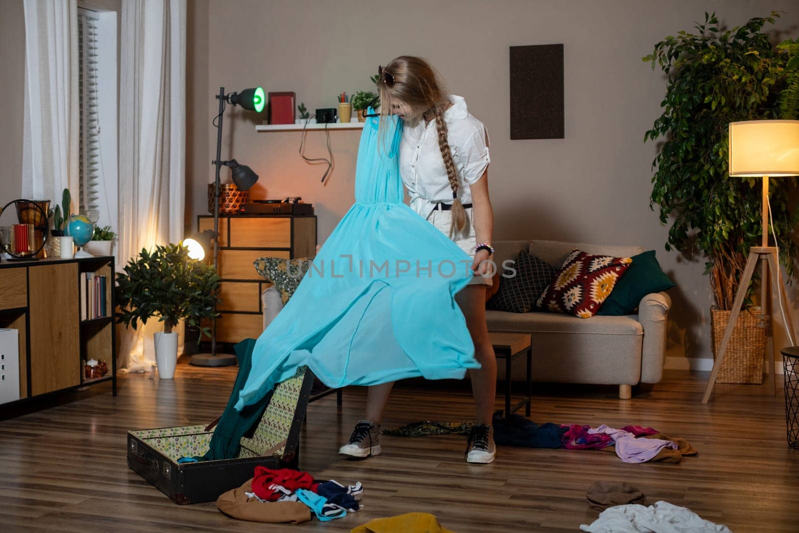 A woman is in a messy room as she sorts through a variety of colorful clothes that she has brought from her suitcase