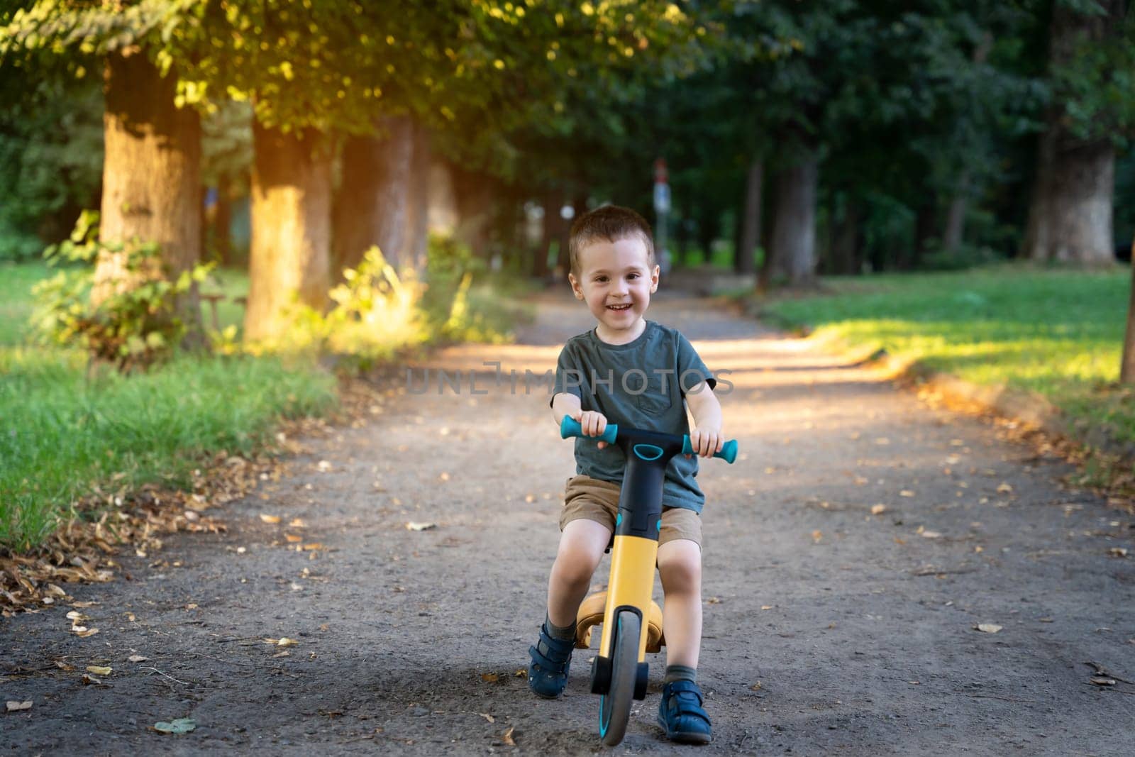 A happy smiling toddler boy of two or three years old rides a bicycle or balance bike in a city park on a sunny summer day. Toddlerhood and childhood concept. Selective focus